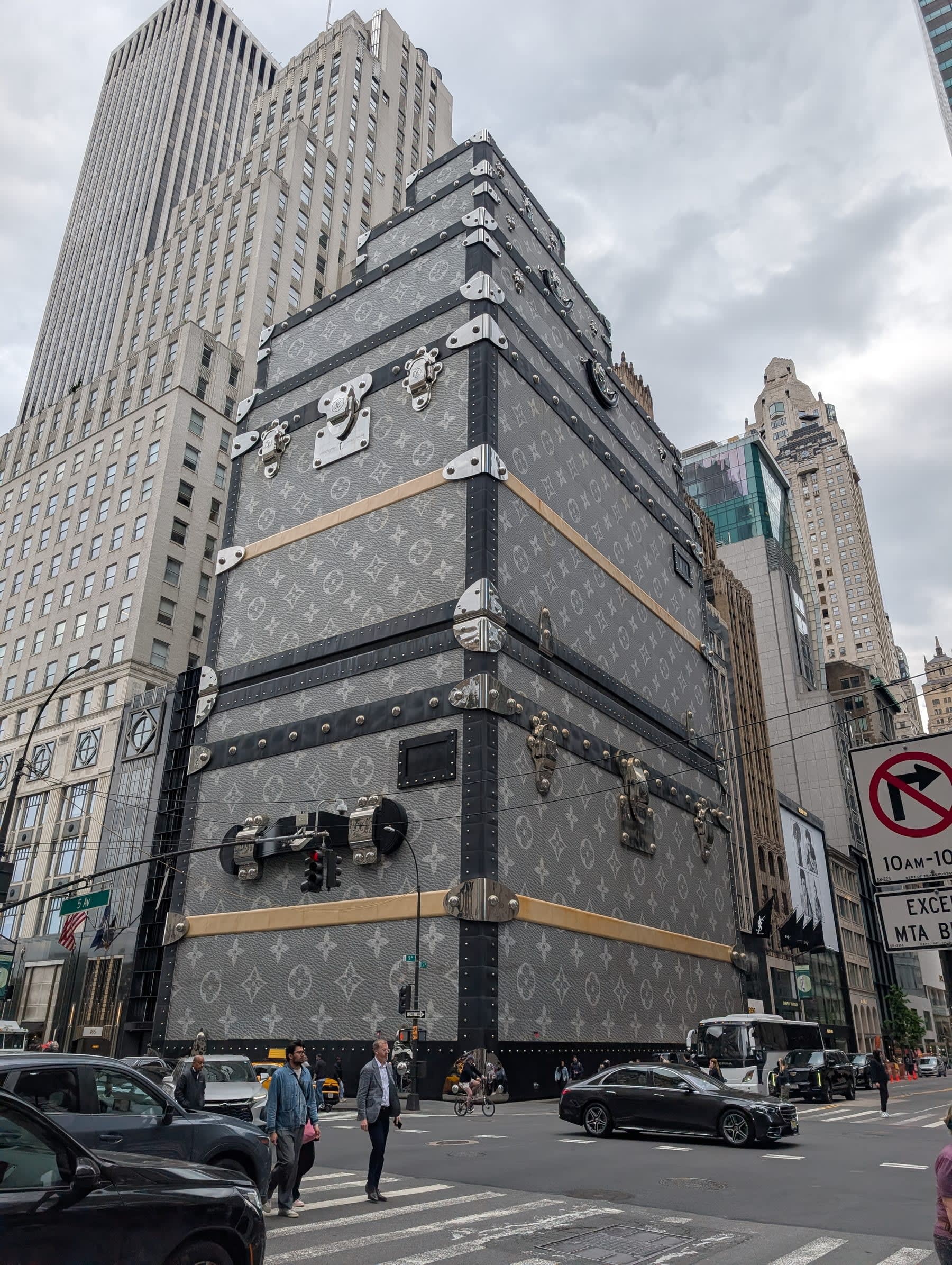 The Louis Vuitton maison on Fifth Avenue — the entire building facade wrapped to resemble stacked trunks in LV monogram canvas, brass hardware at enormous scale, pedestrians and yellow cabs below, the Sherry-Netherland and other midtown towers rising behind it