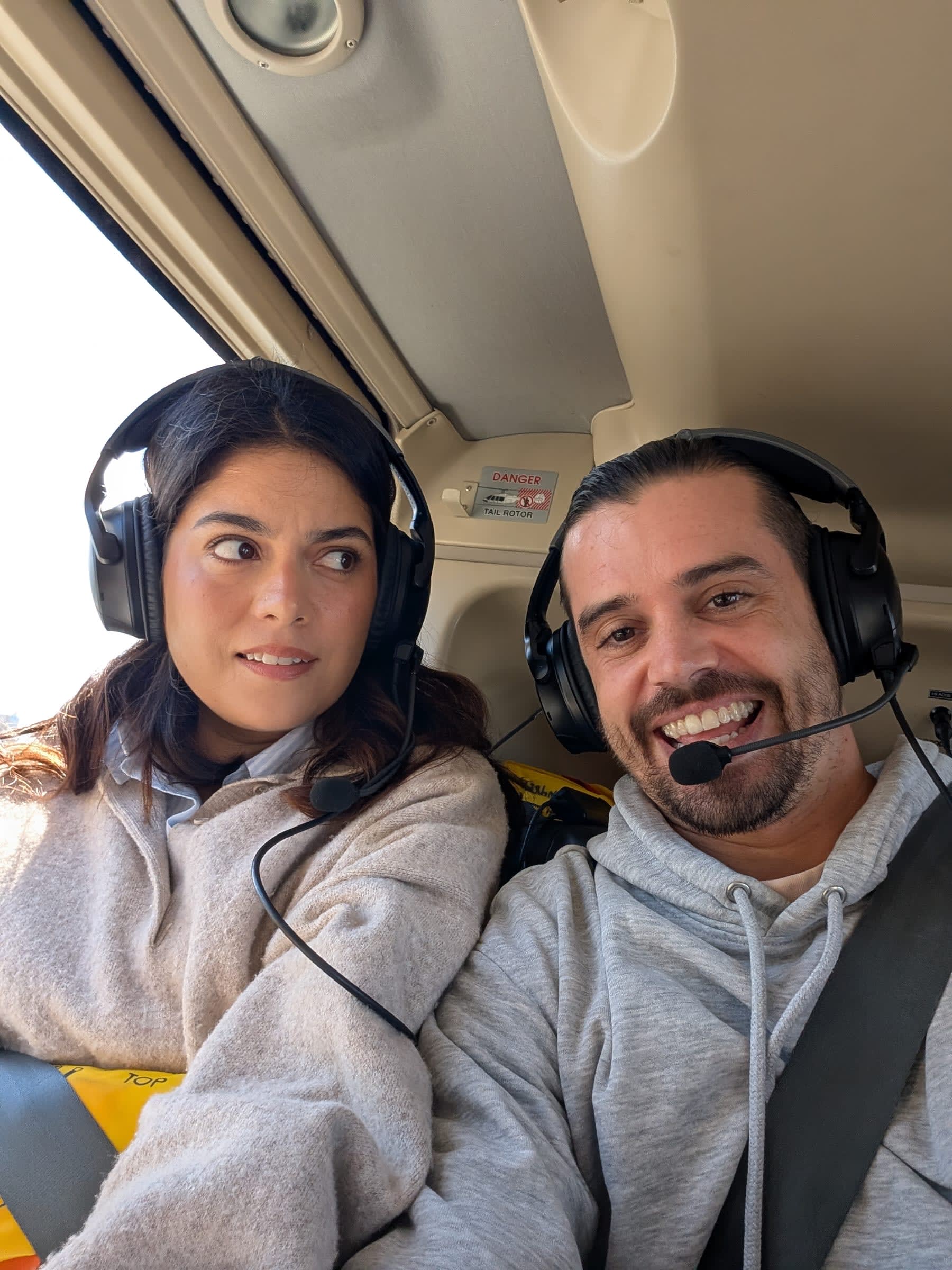 Lia and Pierre in the helicopter cabin wearing aviation headsets — Lia with a sideways look that could be nerves or disbelief, Pierre with the expression of someone who is going to enjoy this regardless