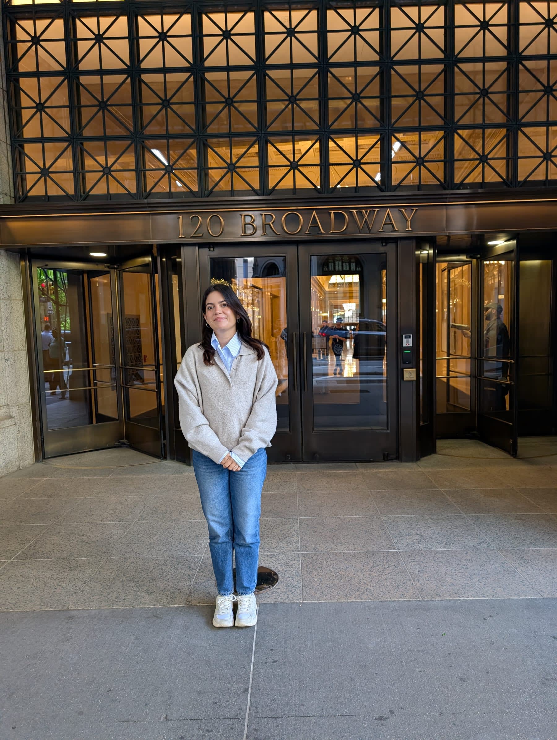 Lia standing in front of the 120 Broadway entrance — gold lettering above the revolving door, an ornate Art Deco entrance canopy, the tower rising out of frame above her