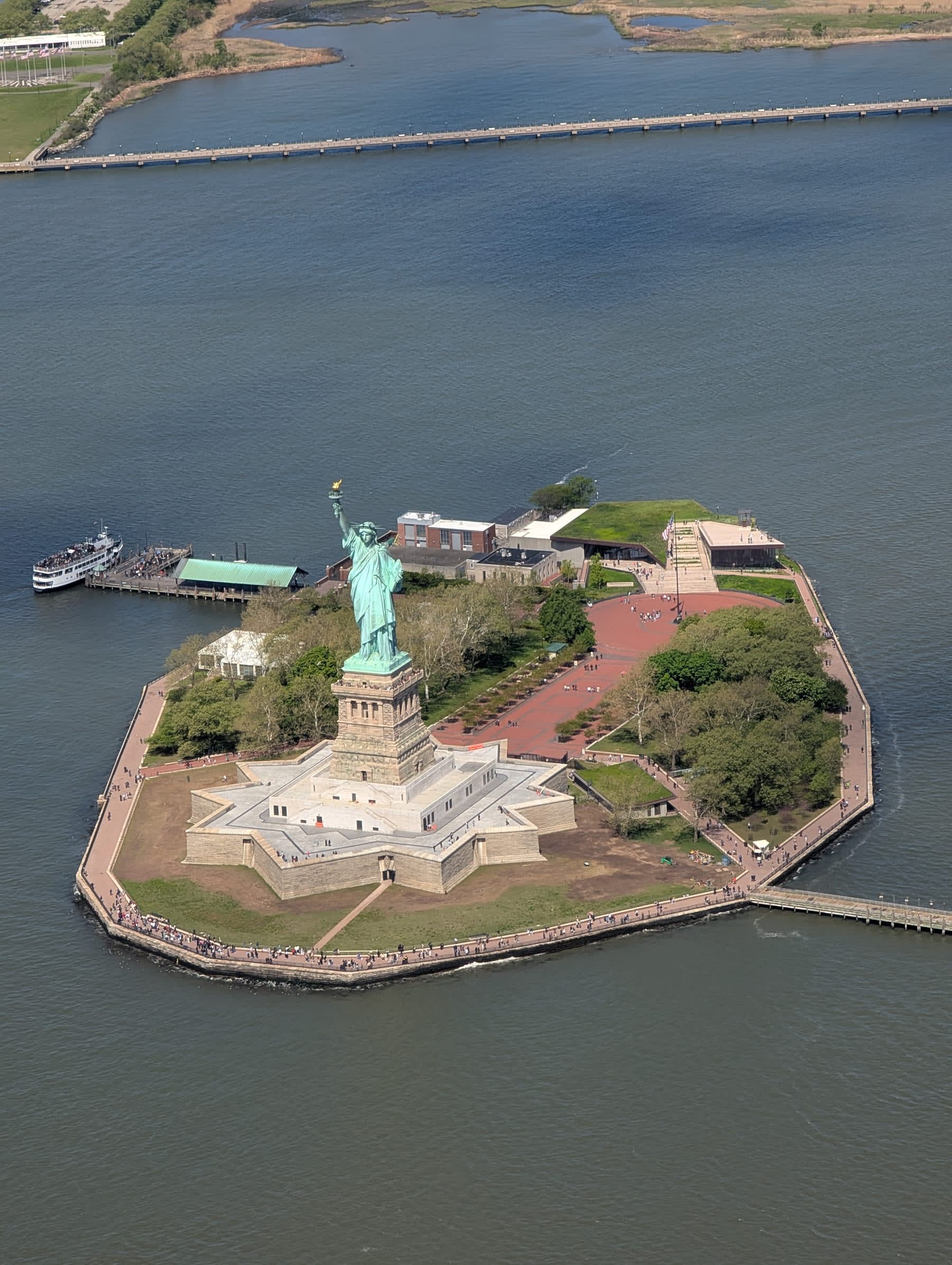 Bird's-eye view of Liberty Island — the Statue of Liberty at the centre, the star-shaped fortifications visible, the ferry dock, the green of the park, surrounded on all sides by the grey-green harbor water