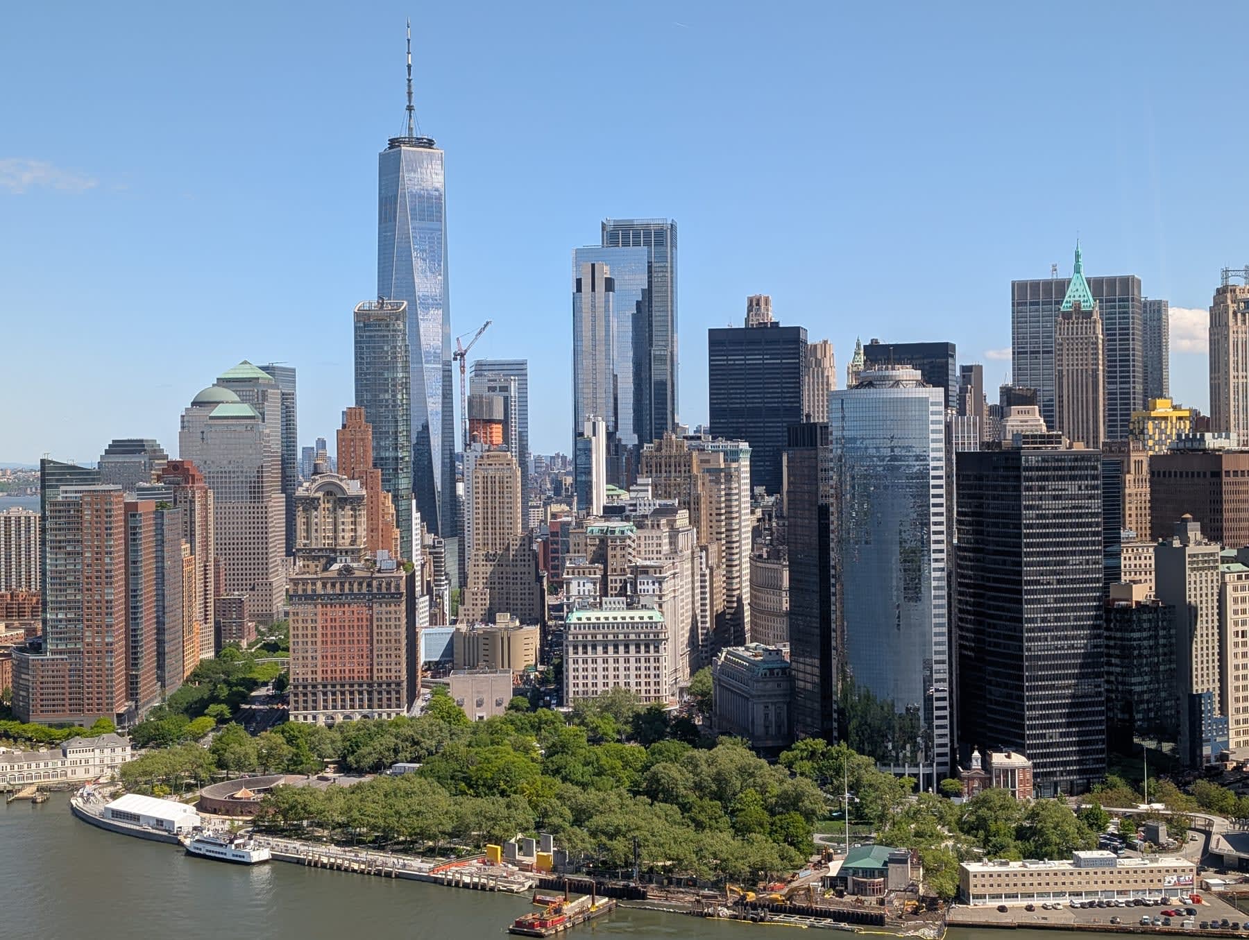 Aerial view of Lower Manhattan from the Hudson — One World Trade Centre dominating the skyline, the older FiDi towers clustered around it, the Battery at the island's tip, the East River and Brooklyn Bridge visible at right