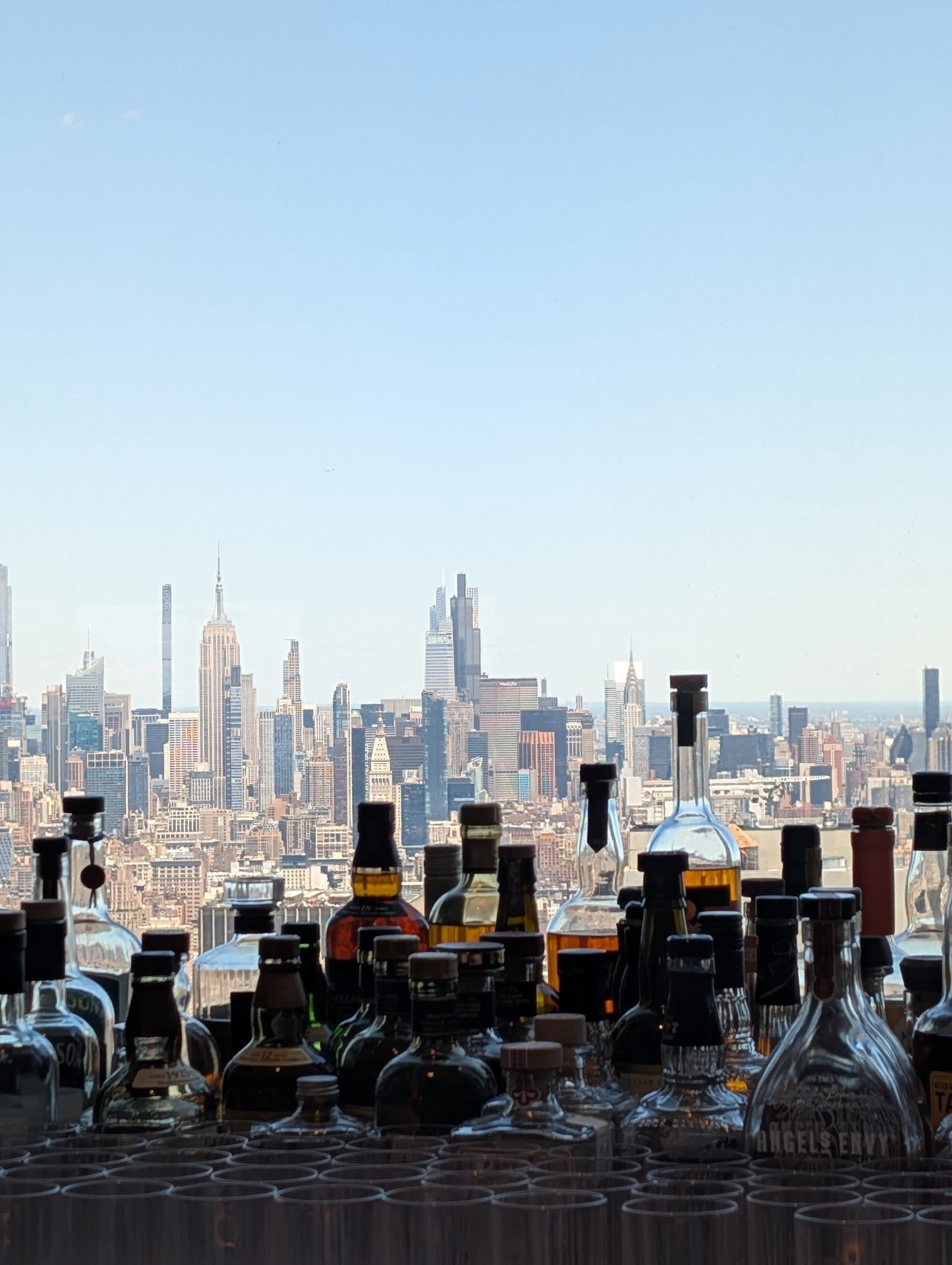 The Manhatta bar — spirits and decanters lined up on a shelf, and beyond them through the floor-to-ceiling window, the full midtown Manhattan skyline: Empire State Building, Chrysler Building, and the midtown cluster floating in pale afternoon light
