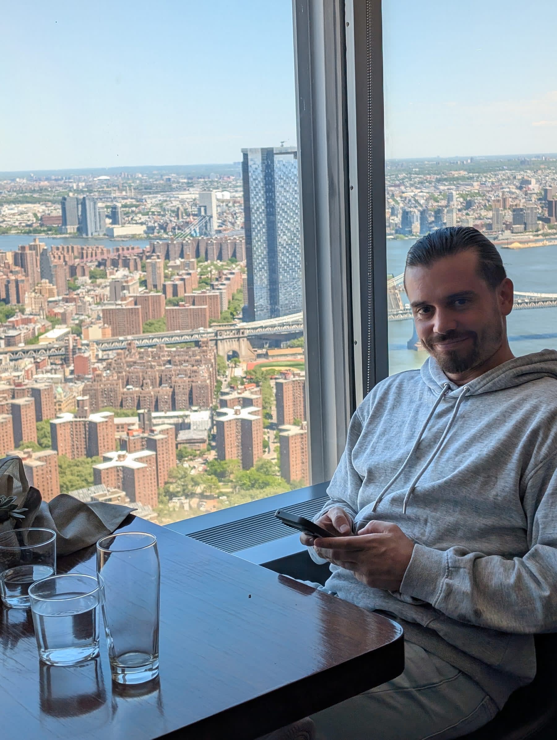 Pierre seated at a window table at Manhatta — phone in hand, relaxed, the vast view of Brooklyn and the East River spread behind him through the floor-to-ceiling glass, the lower Manhattan rooftops far below
