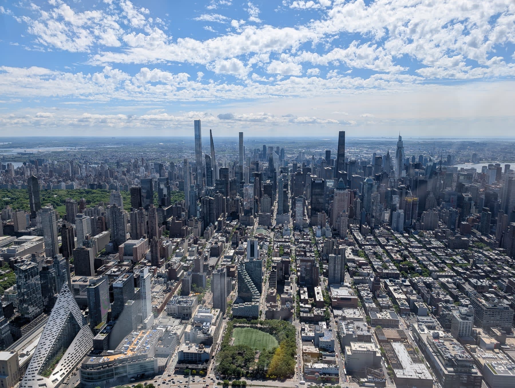 Aerial panoramic looking north over midtown Manhattan — Central Park visible as a perfect green rectangle, the Steinway Tower and 432 Park Avenue rising needle-thin above the dense grid, the Hudson and East River flanking the island on both sides