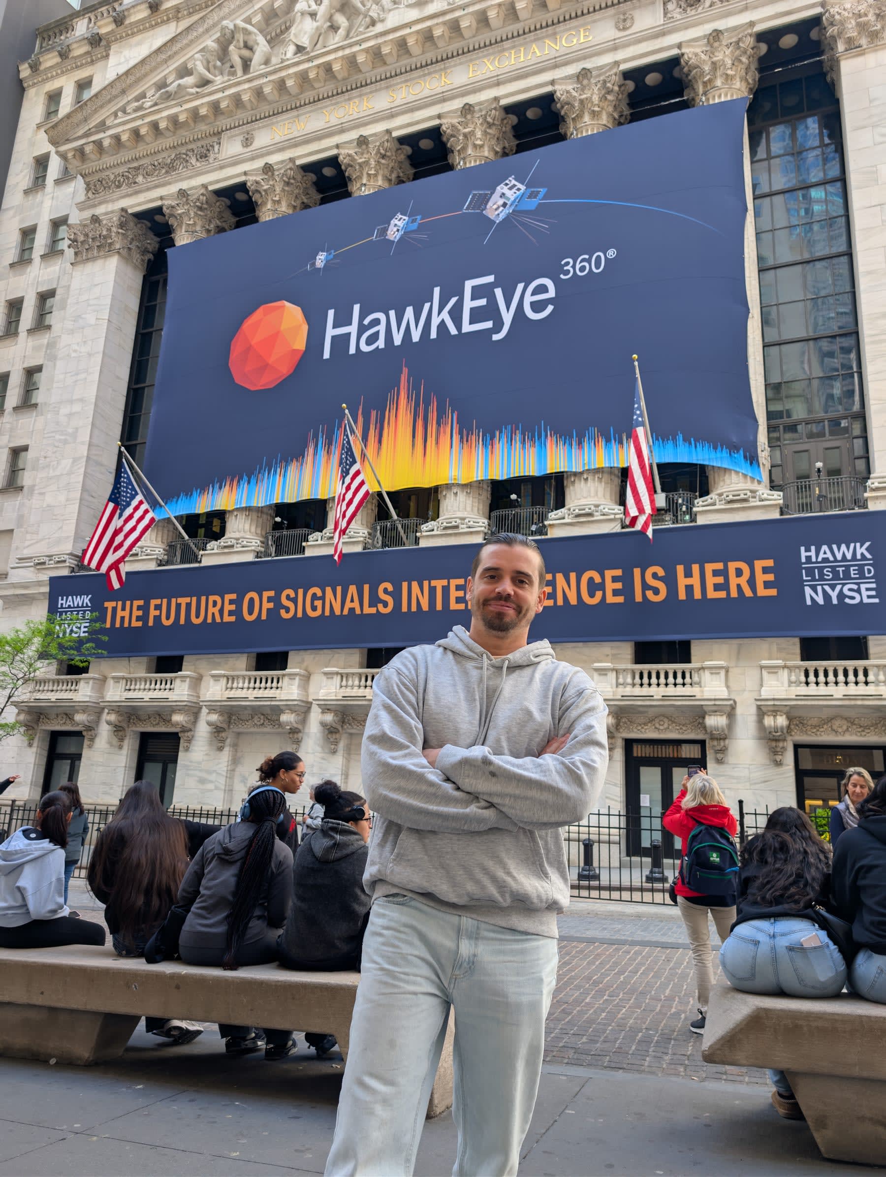 Pierre standing with arms folded in front of the New York Stock Exchange — the Corinthian columns behind him, a large HawkEye 360 banner covering the upper facade, American flags on either side