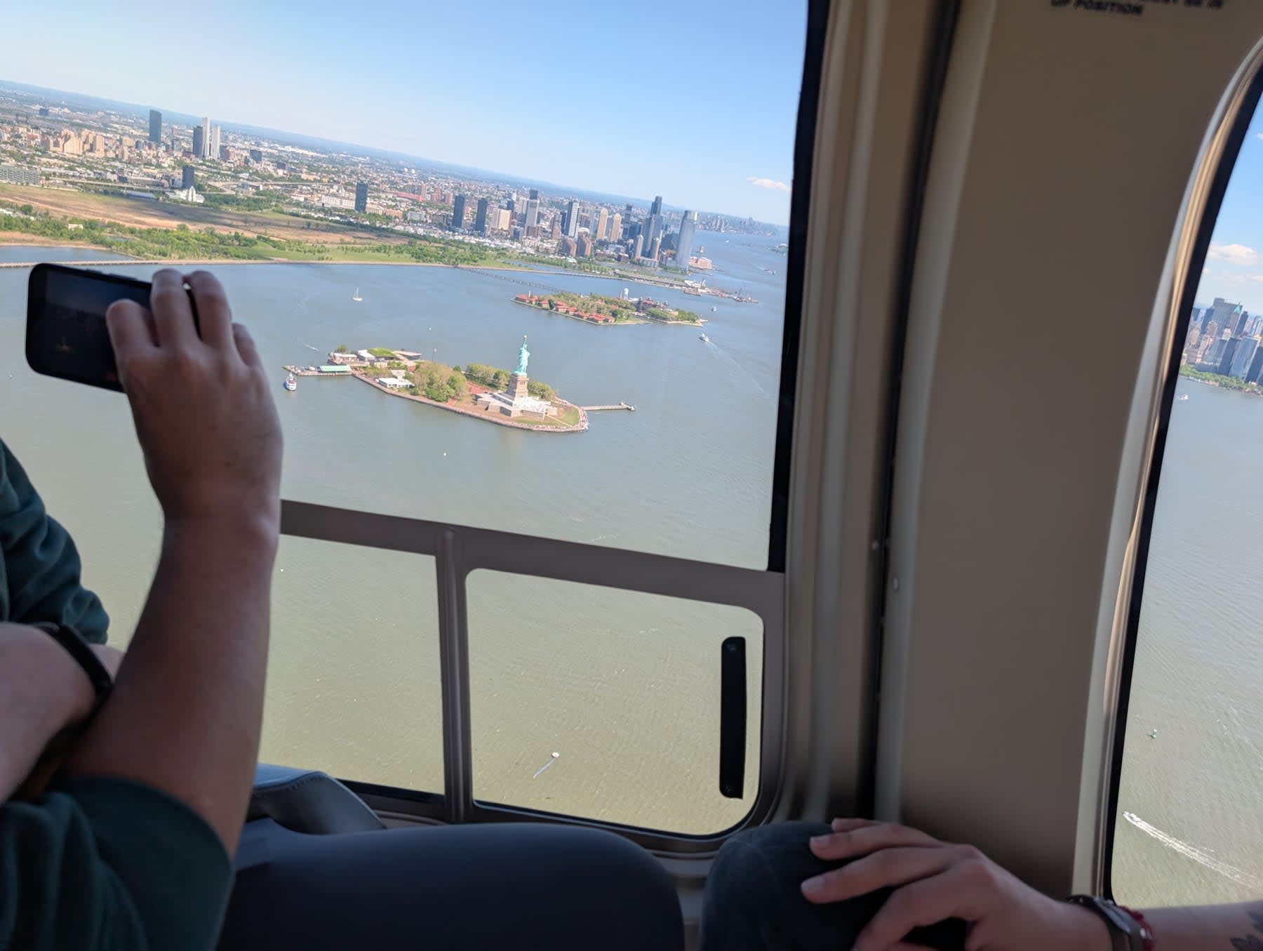 View through the helicopter window — someone holding a phone to photograph the Statue of Liberty far below, Liberty Island small in the harbor, New Jersey stretching behind it, the tip of Manhattan visible at right