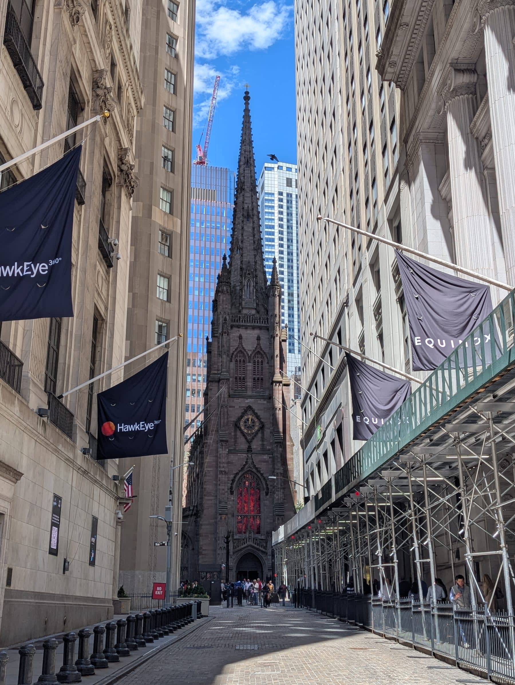 Wall Street looking west toward Broadway — tall stone and glass buildings forming a canyon on both sides, HawkEye banners hanging from the NYSE at left, Trinity Church's Gothic spire framed at the end of the corridor under blue sky