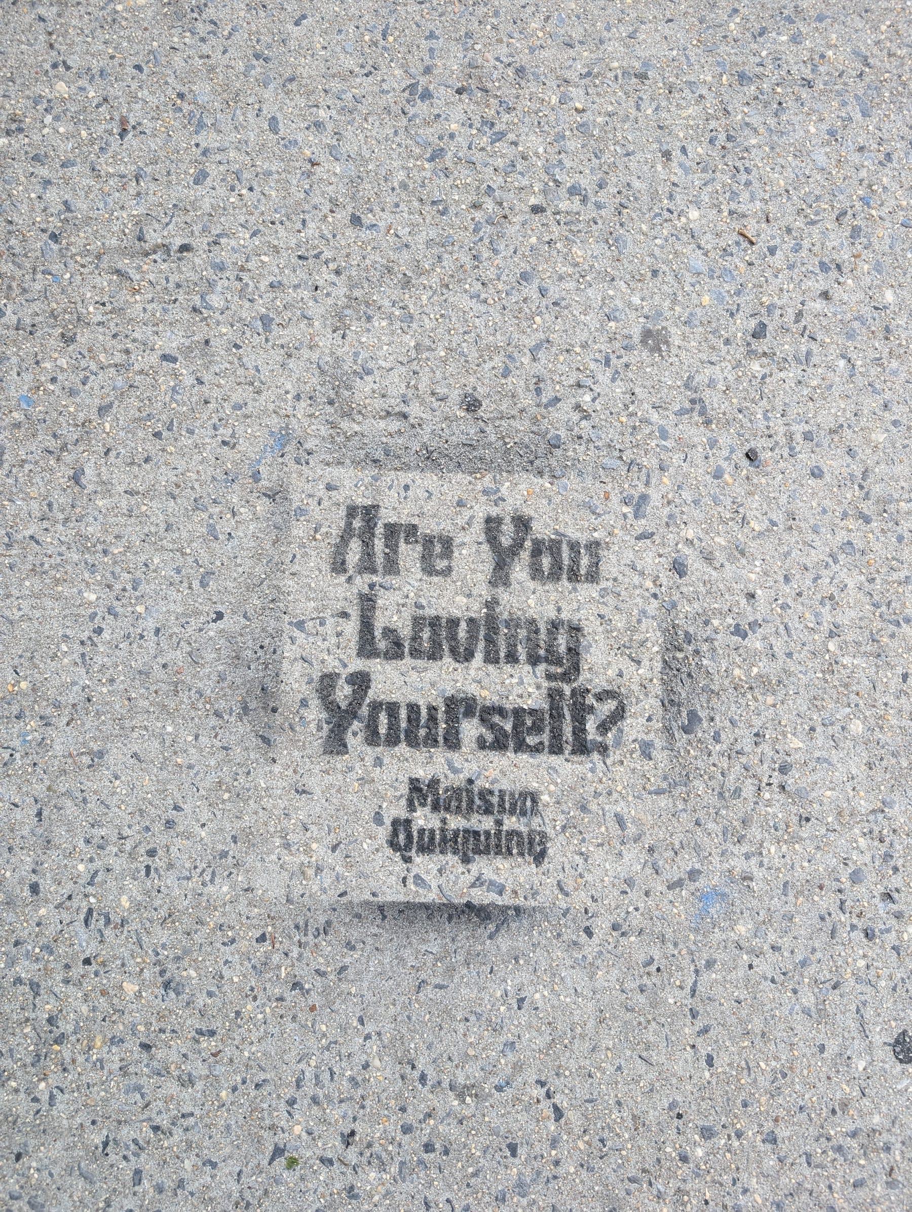 A concrete sidewalk stencilled with "Are You Loving Yourself?" in bold black letters, with the brand name "Maison Quinqine" printed below — the city's peculiar form of philosophy, delivered underfoot