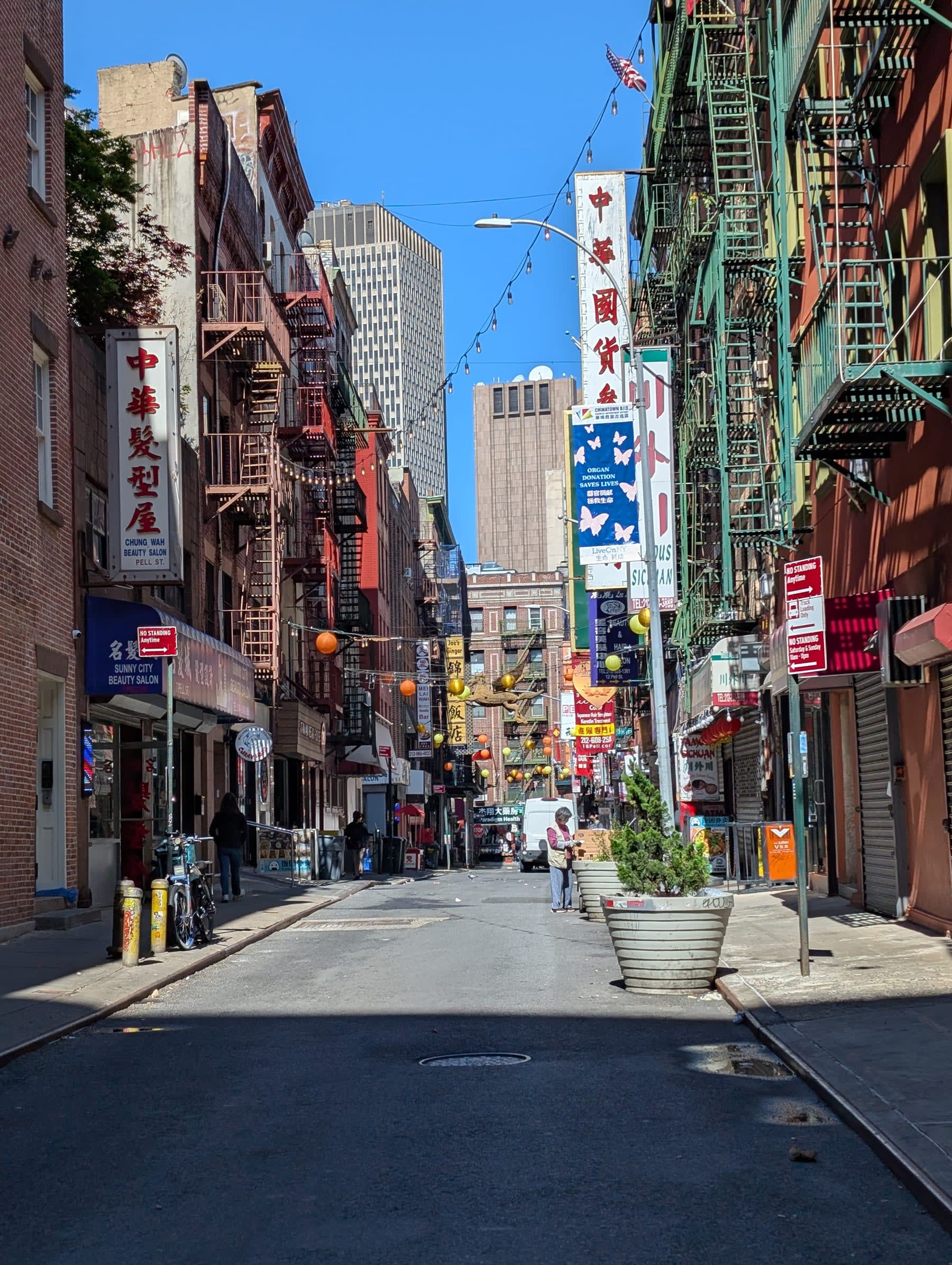 A narrow Chinatown street on a sunny morning — Chinese-language signs and red lanterns on low-rise buildings, a few people on the empty pavement, Financial District towers visible at the far end framed like a painting