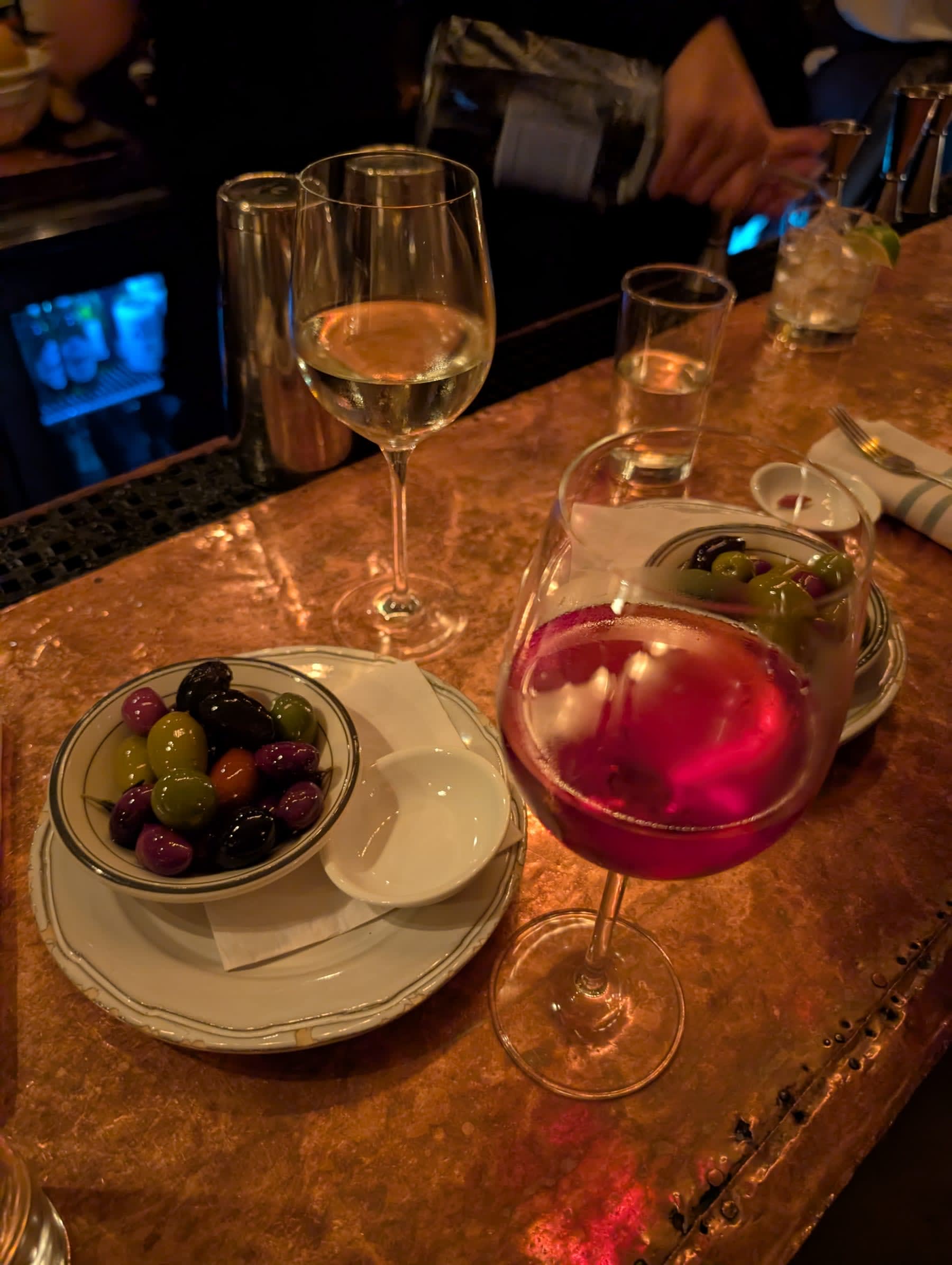 A copper bar counter at close to 2am — a pink cocktail with a citrus garnish, a wine glass of white wine, and a small bowl of mixed olives arranged on the gleaming surface under warm light