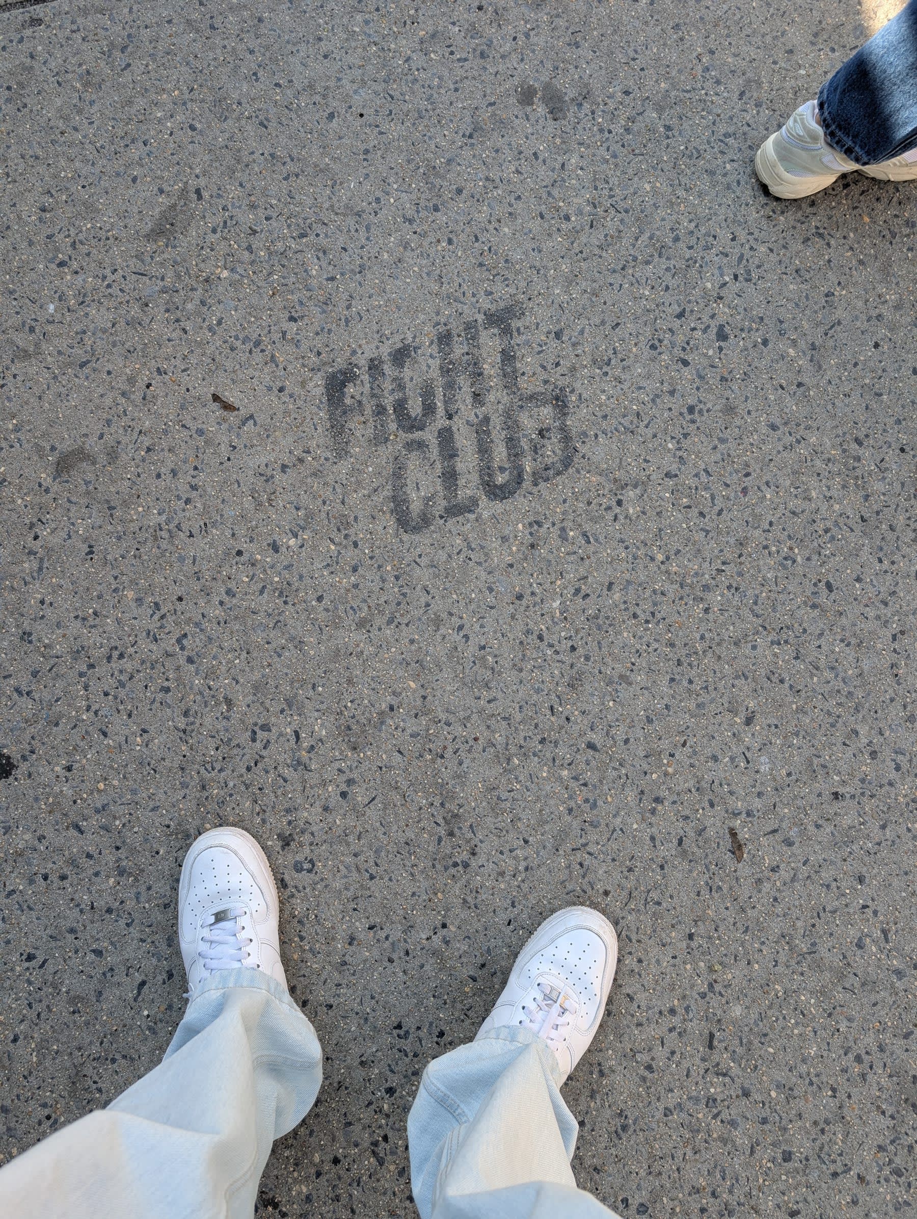 Pierre's white sneakers on a concrete pavement, the words "FIGHT CLUB" stencilled in large black letters between his feet — Lia's jeans and shoes visible at the top right corner