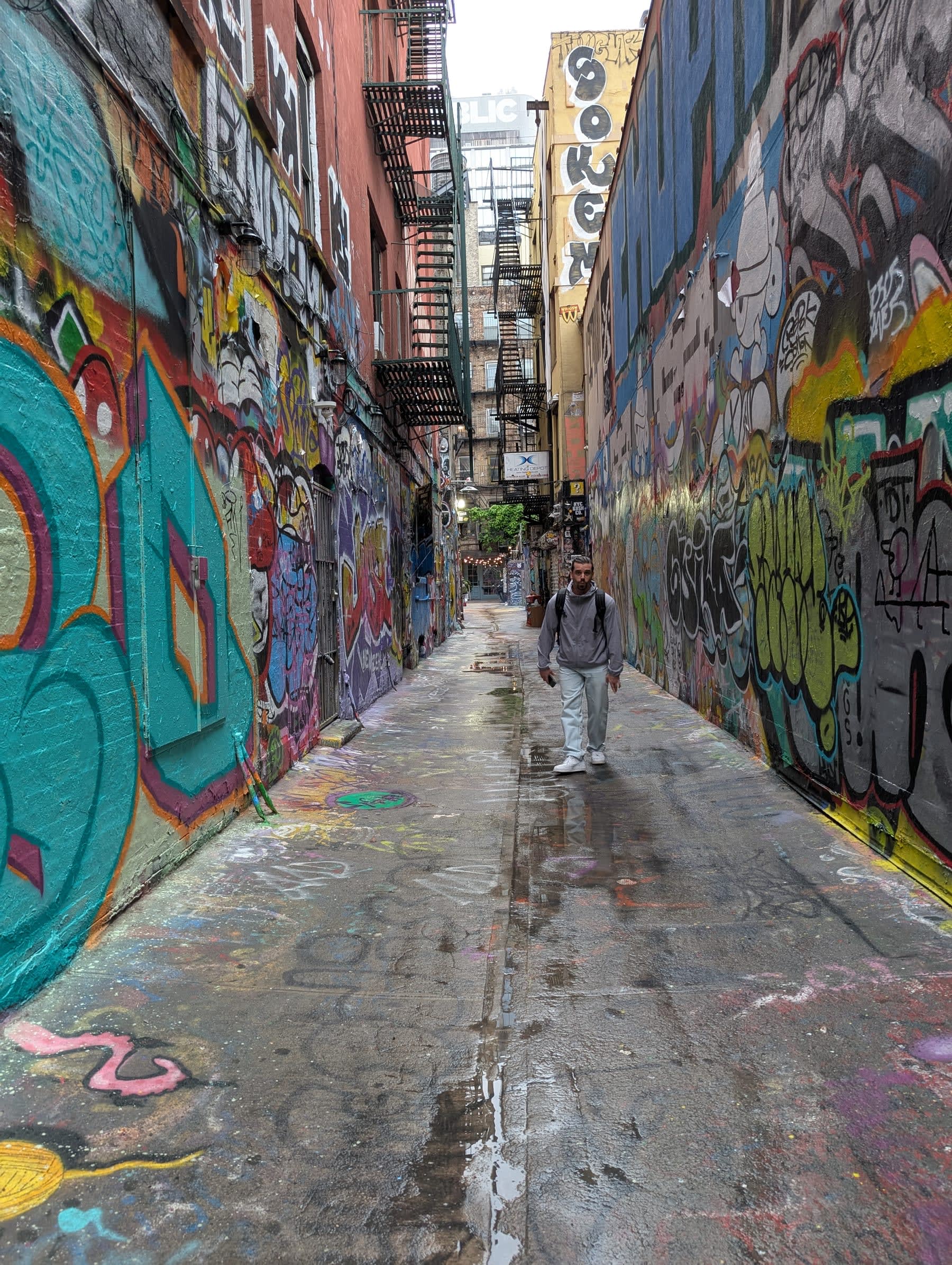 Pierre walking through Freeman Alley — graffiti-covered walls rising four storeys on both sides, fire escapes layered overhead, the alley narrowing toward a warm light at the far end