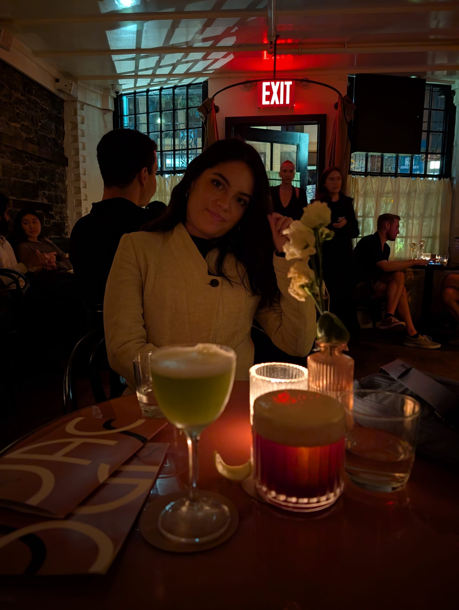 Lia at a candlelit table inside Freemans at midnight — a green cocktail and an amber cocktail in front of her, a white flower in a small vase, the low dark-panelled room and other tables visible behind her