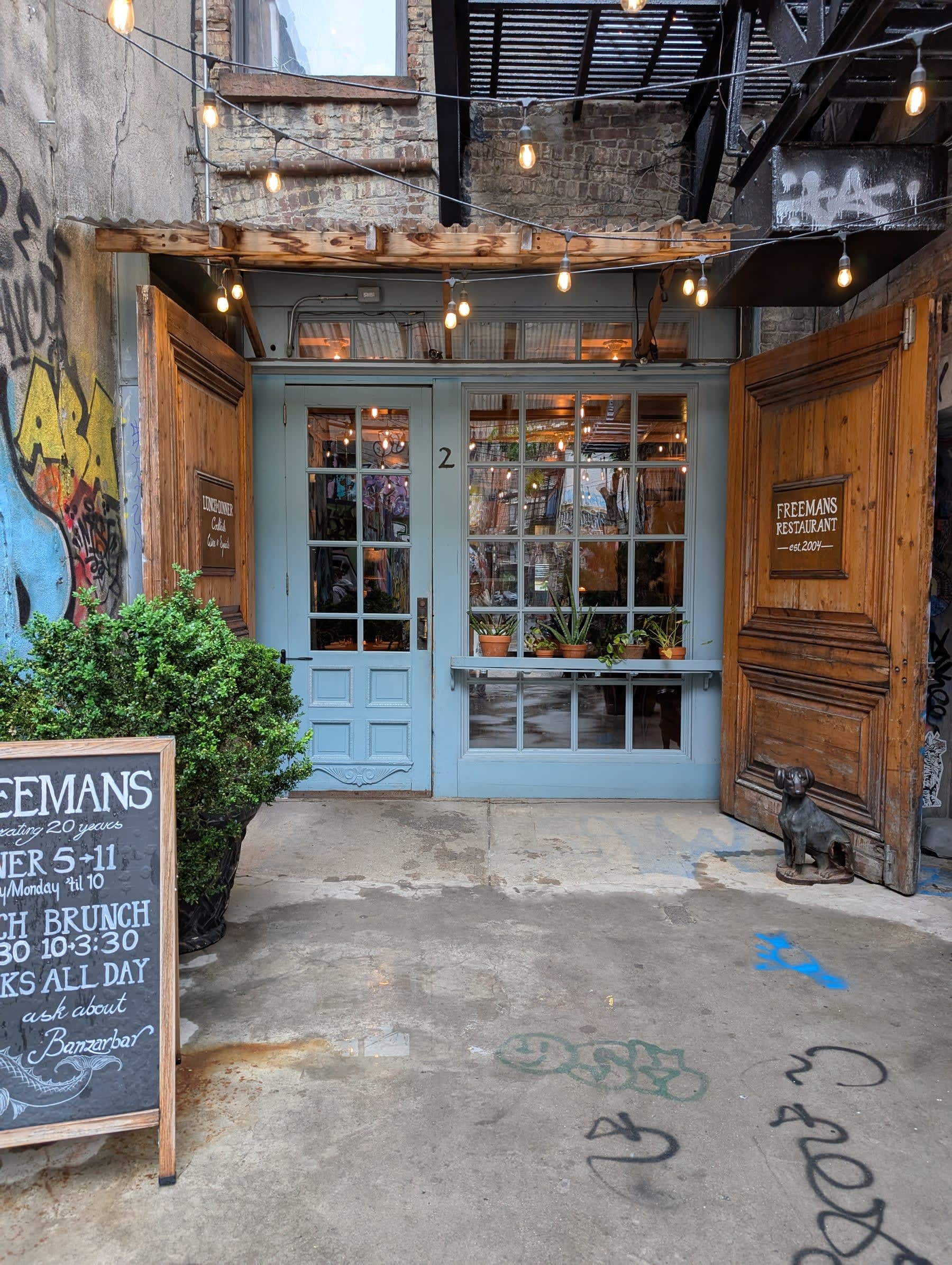 The entrance to Freemans restaurant — a pale blue painted door with small-paned windows, Edison bulbs strung overhead, a handwritten chalkboard listing the hours beside an iron dog figurine
