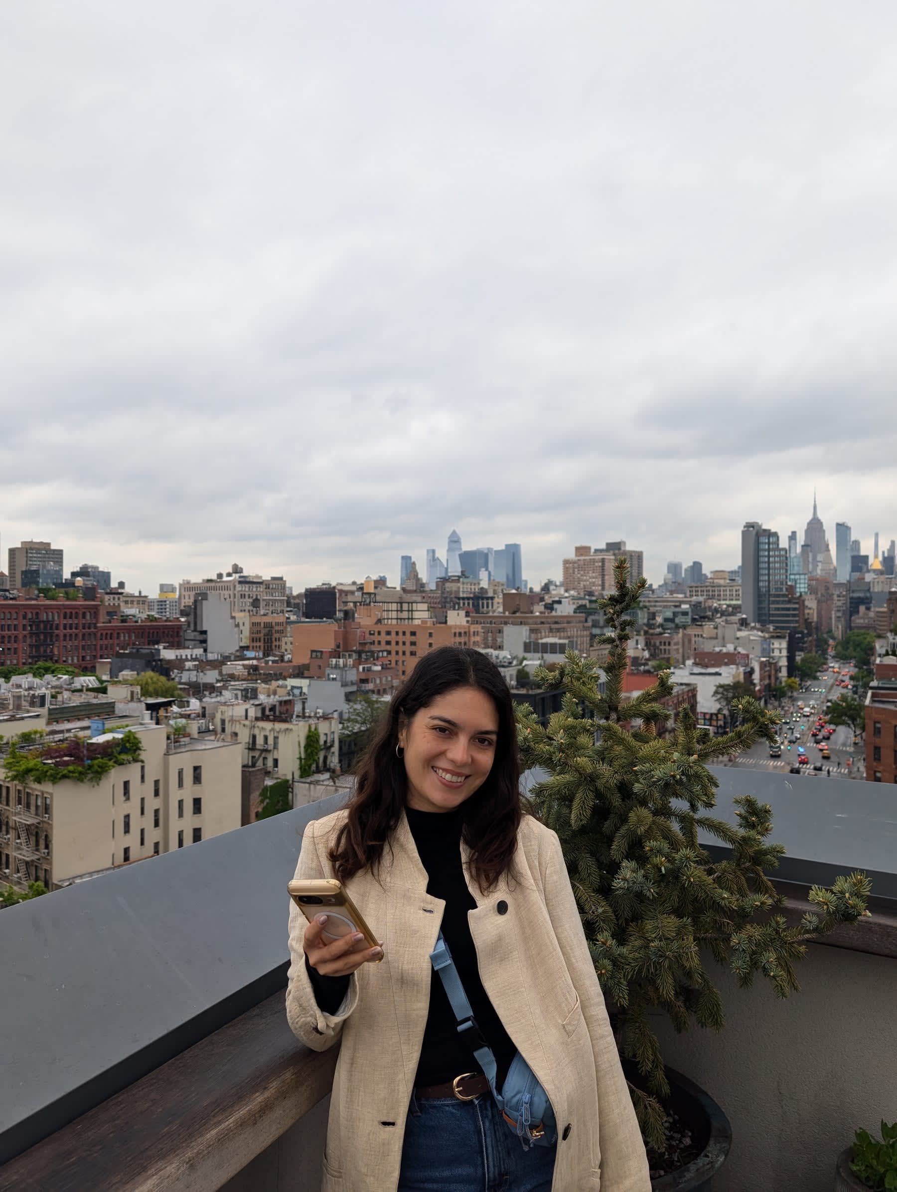 Lia on the hotel rooftop smiling at the camera, the full Manhattan skyline — Empire State Building, midtown towers — stretching across the grey May sky behind her