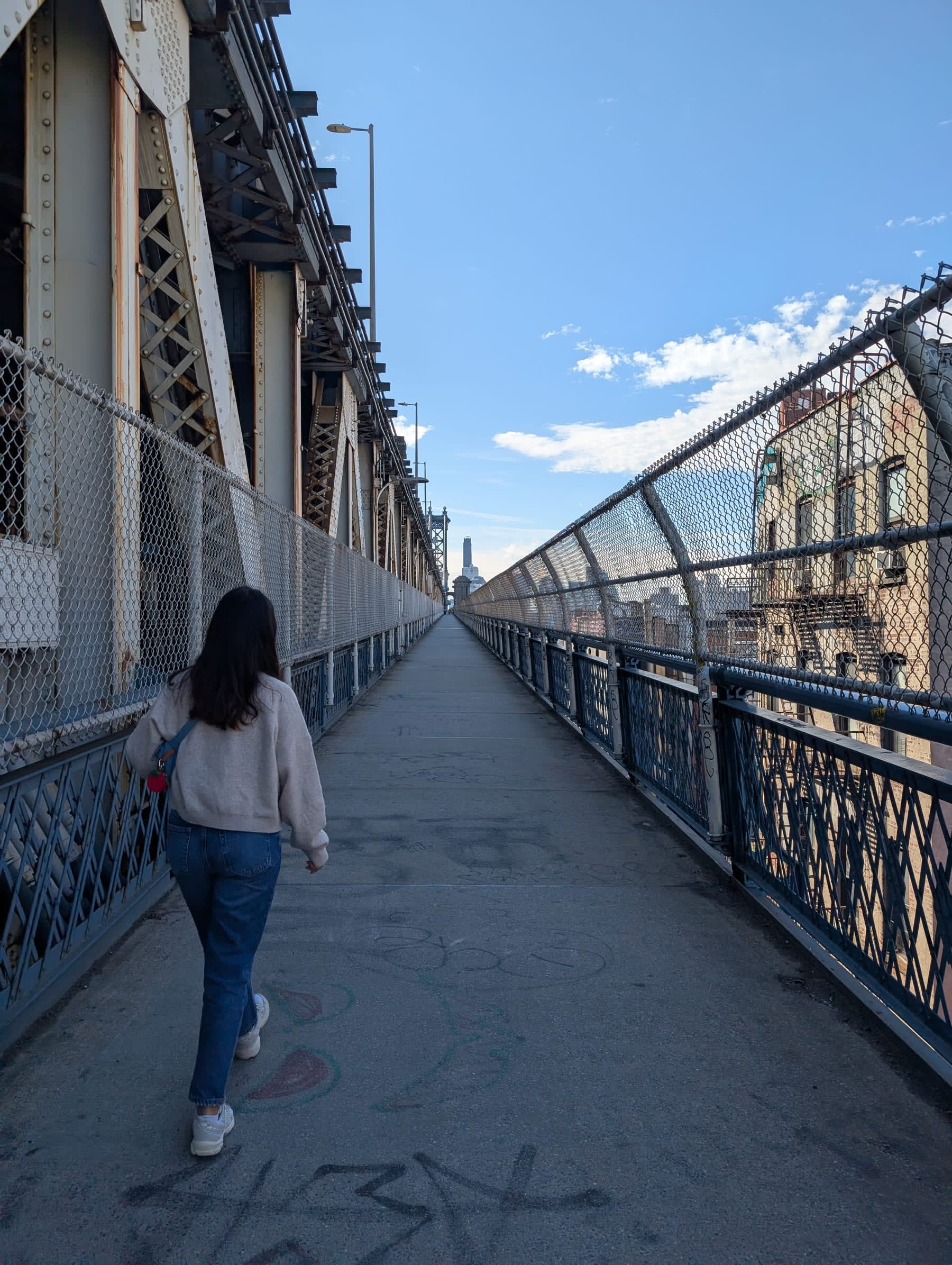Lia walking away down the Manhattan Bridge pedestrian walkway — blue sky above, steel lattice and chain-link fencing on both sides, the walkway stretching ahead toward Brooklyn