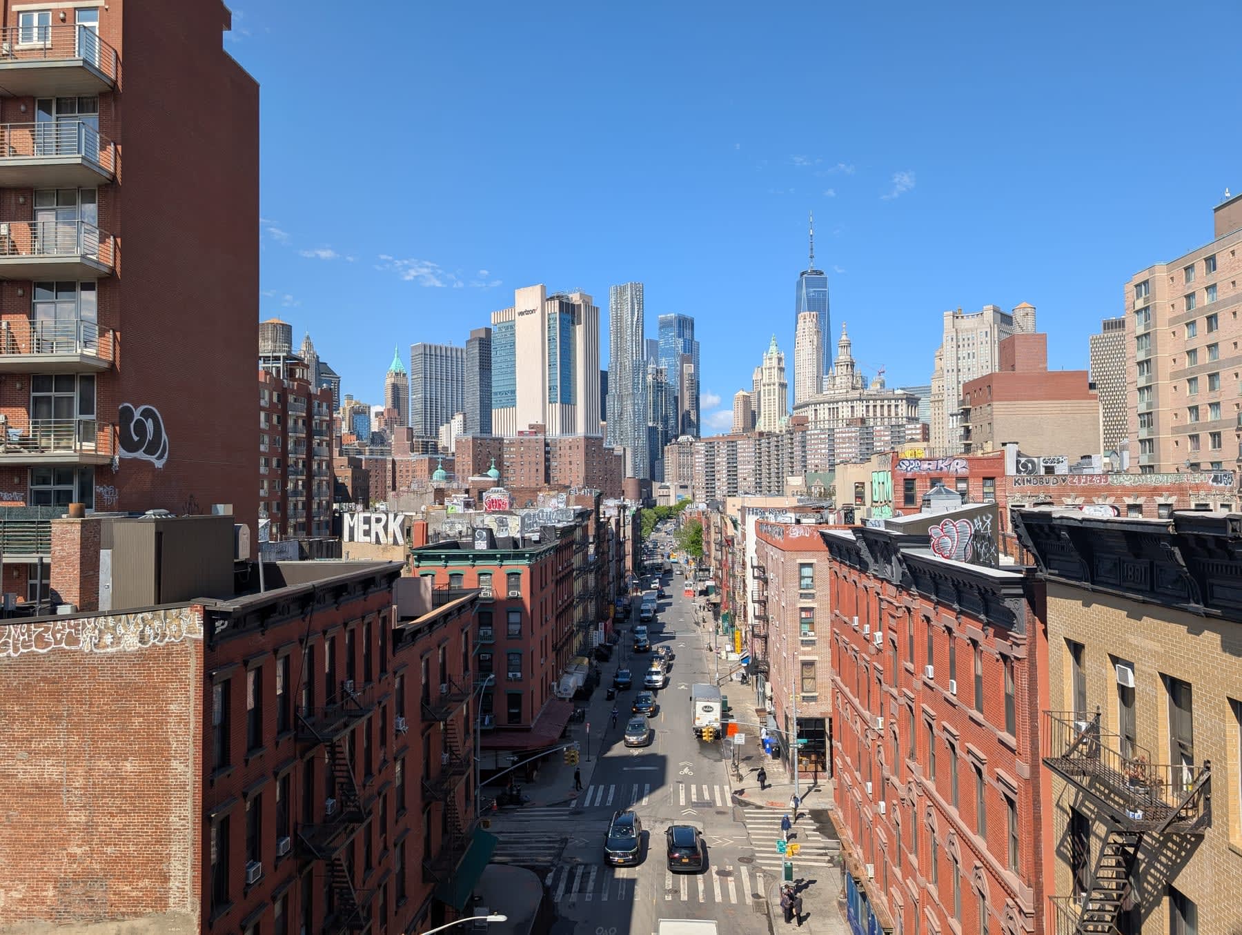 Looking south down a Lower East Side street from an elevated vantage — brick tenements, a rooftop tagged with "MERK", One World Trade Centre visible at the end of the corridor under clear blue sky