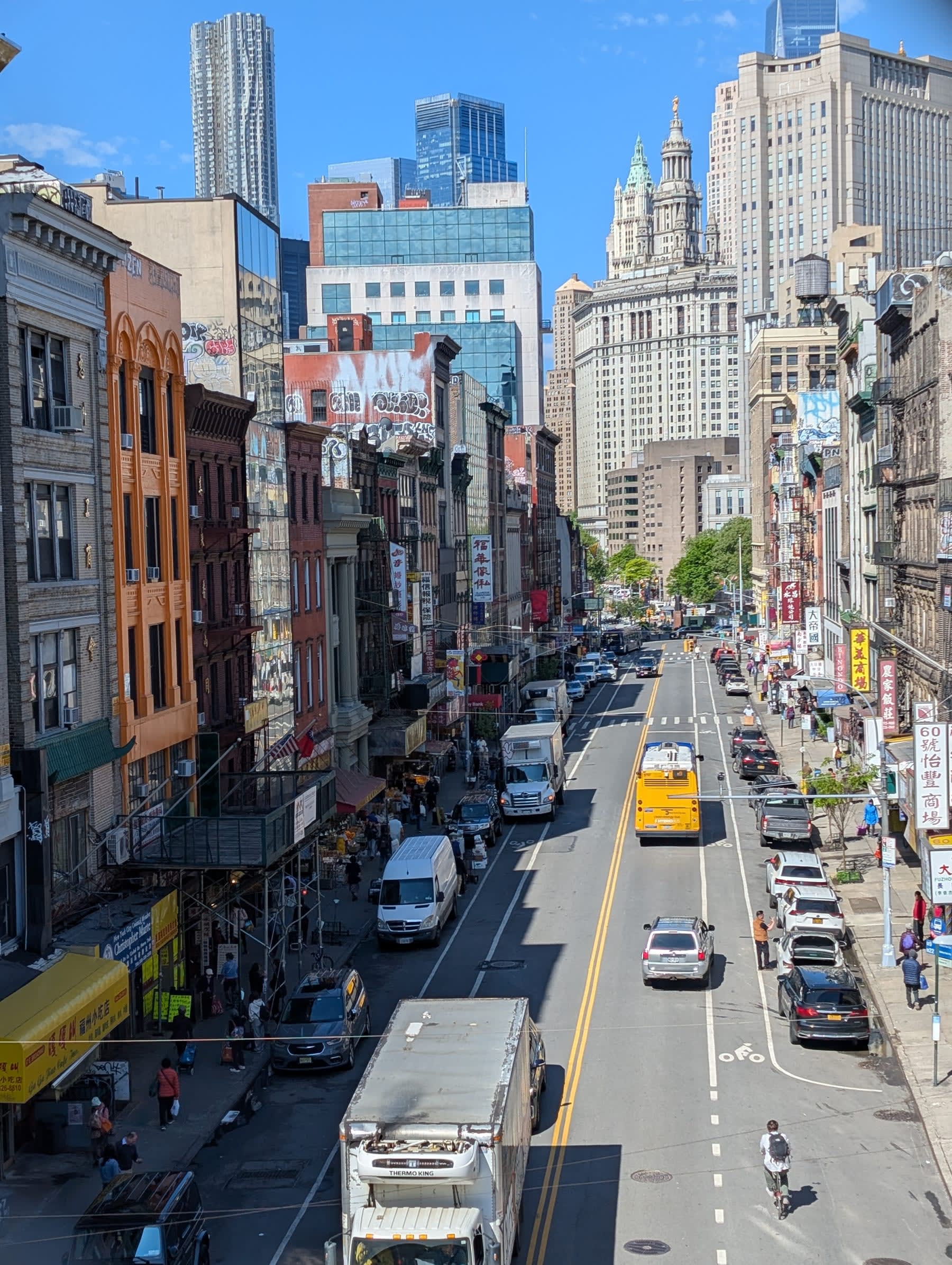Looking back toward Manhattan from the elevated Manhattan Bridge walkway — a long avenue cutting through Chinatown below, the Municipal Building dome and Financial District towers rising in the background under blue sky