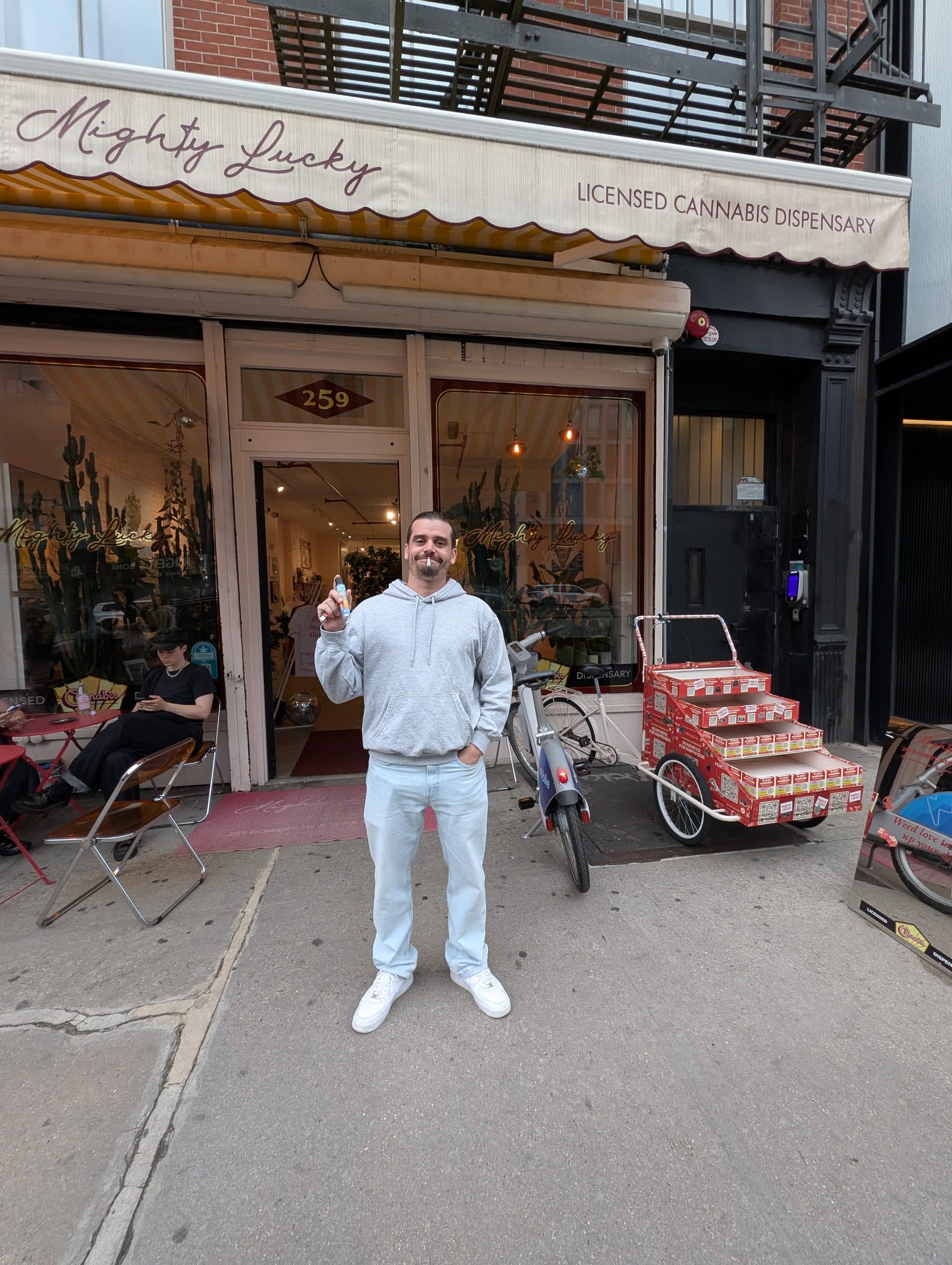 Pierre standing in front of Mighty Lucky at 259 — a licensed cannabis dispensary with a cream awning, giving a thumbs up, a cargo bike stacked with cases parked beside him on the sidewalk