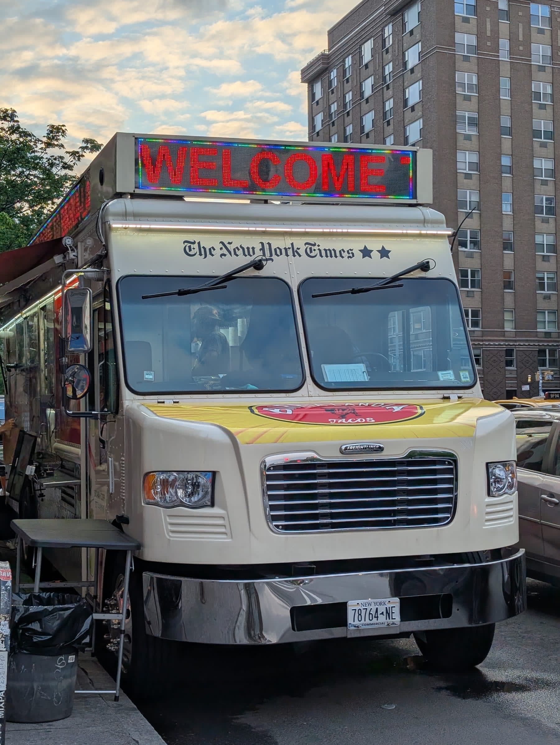 A New York Times-branded food truck parked on a quiet side street at night, the LED marquee sign on top reading "WELCOME" in red block letters, street lights visible in the background