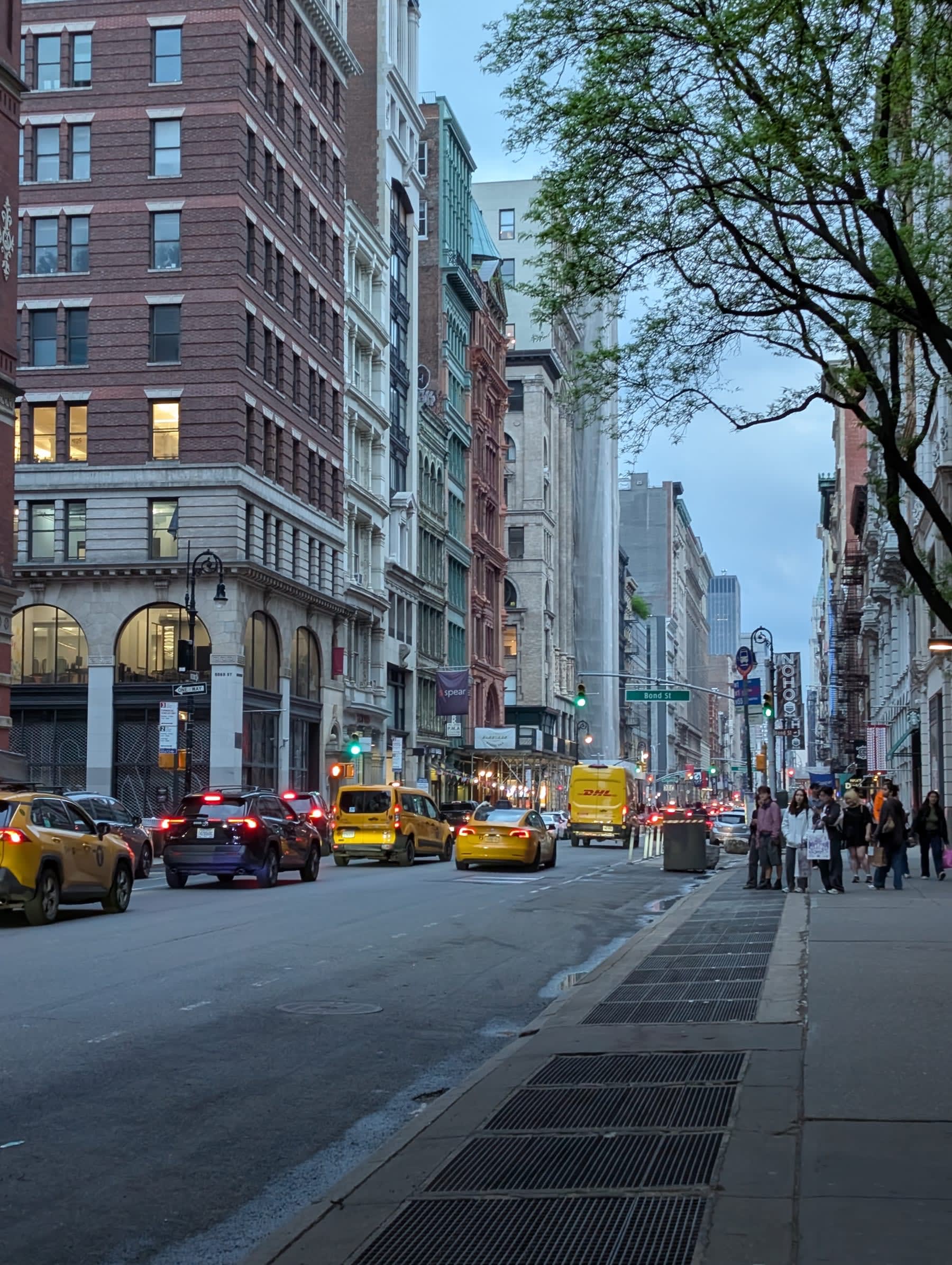 A long SoHo avenue at blue hour — yellow cabs in the intersection, cast-iron facades and lit storefronts stretching into the distance, a green traffic light and pedestrians beginning to cross