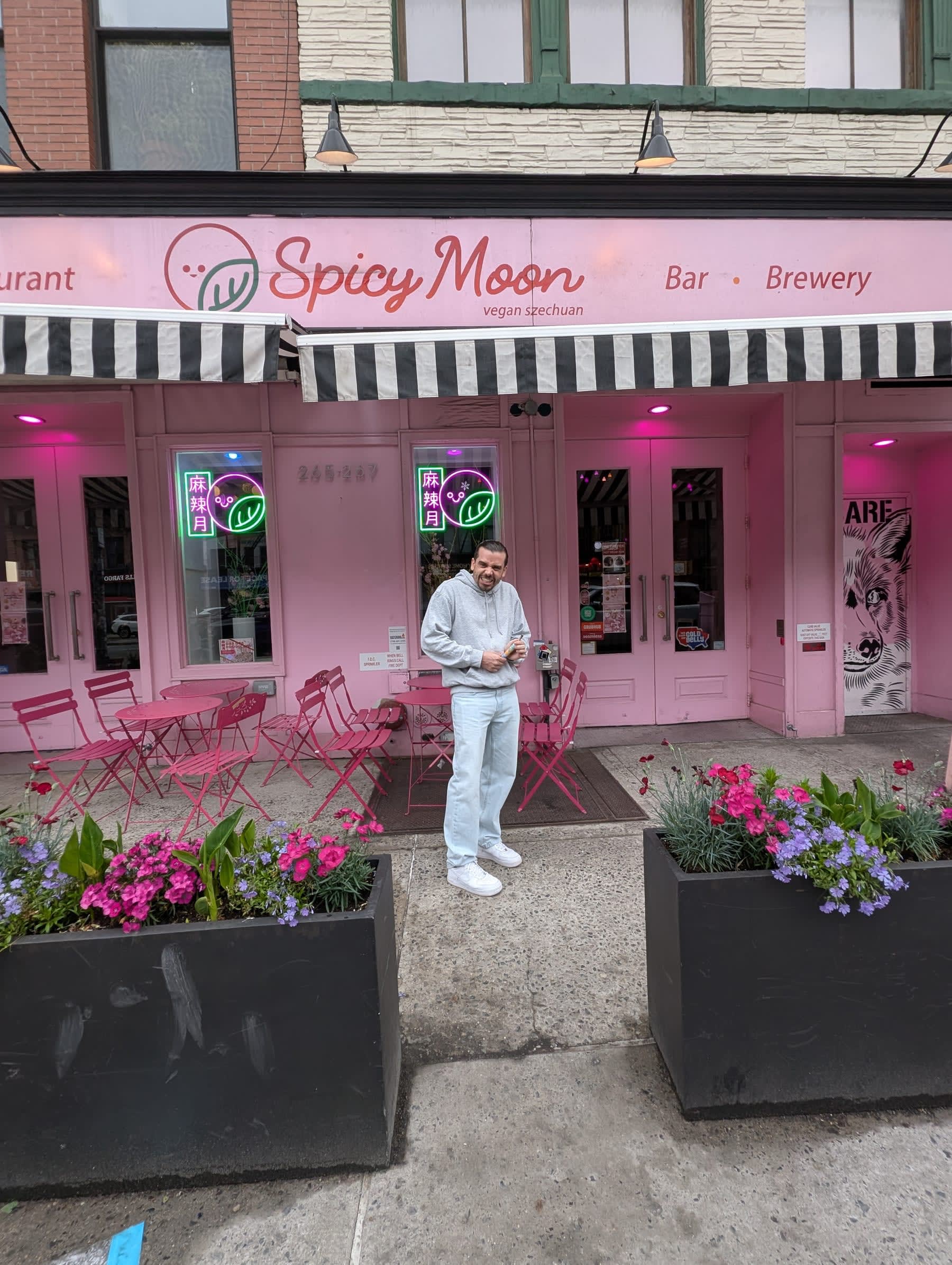 Pierre standing in front of Spicy Moon, a vegan Sichuan restaurant with a bright pink and white exterior, flower planters on both sides of the entrance, the words "Bar · Brewery" beside the logo