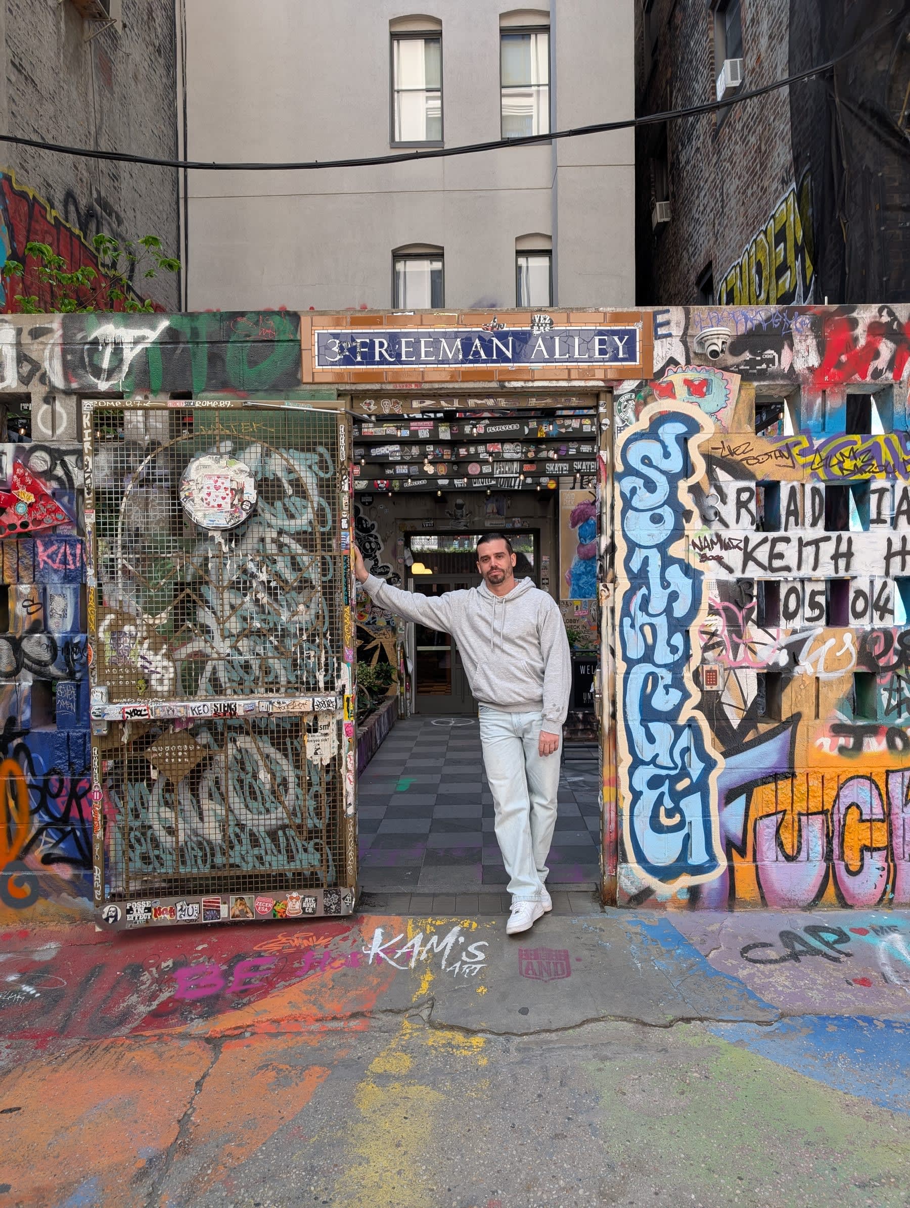 Pierre posing at the entrance gate of Freeman Alley — the "3 Freeman Alley" sign above him, graffiti-covered walls on both sides, leaning casually against the gatepost before an evening out