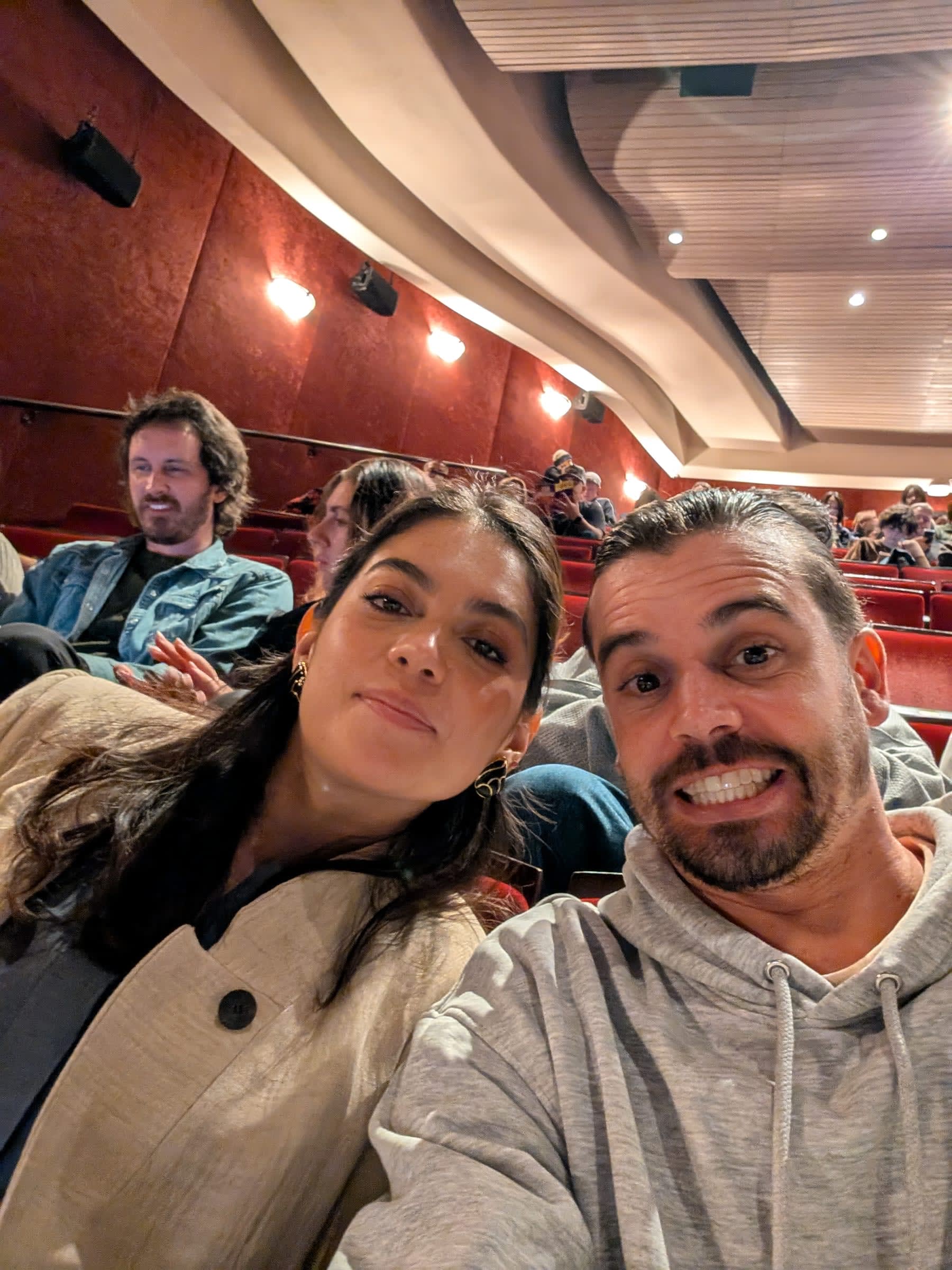 Lia and Pierre selfie in their Marquis Theatre seats — Pierre grinning broadly, Lia composed and smiling, the theatre auditorium and other audience members visible behind them