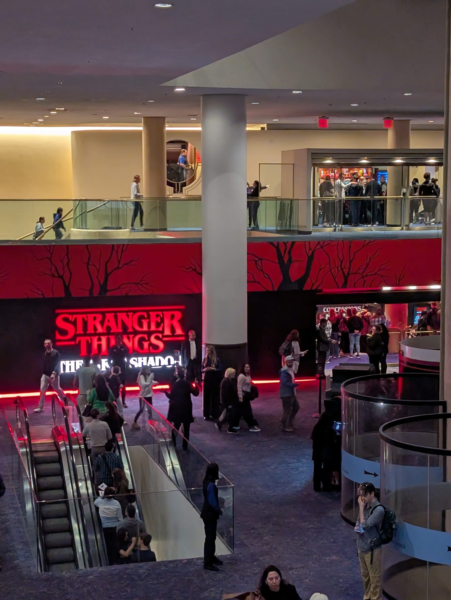 The Marquis Theatre lobby from the mezzanine — the "Stranger Things: The First Shadow" sign glowing in red neon against a black background painted with bare winter trees, audience members filling the lower level