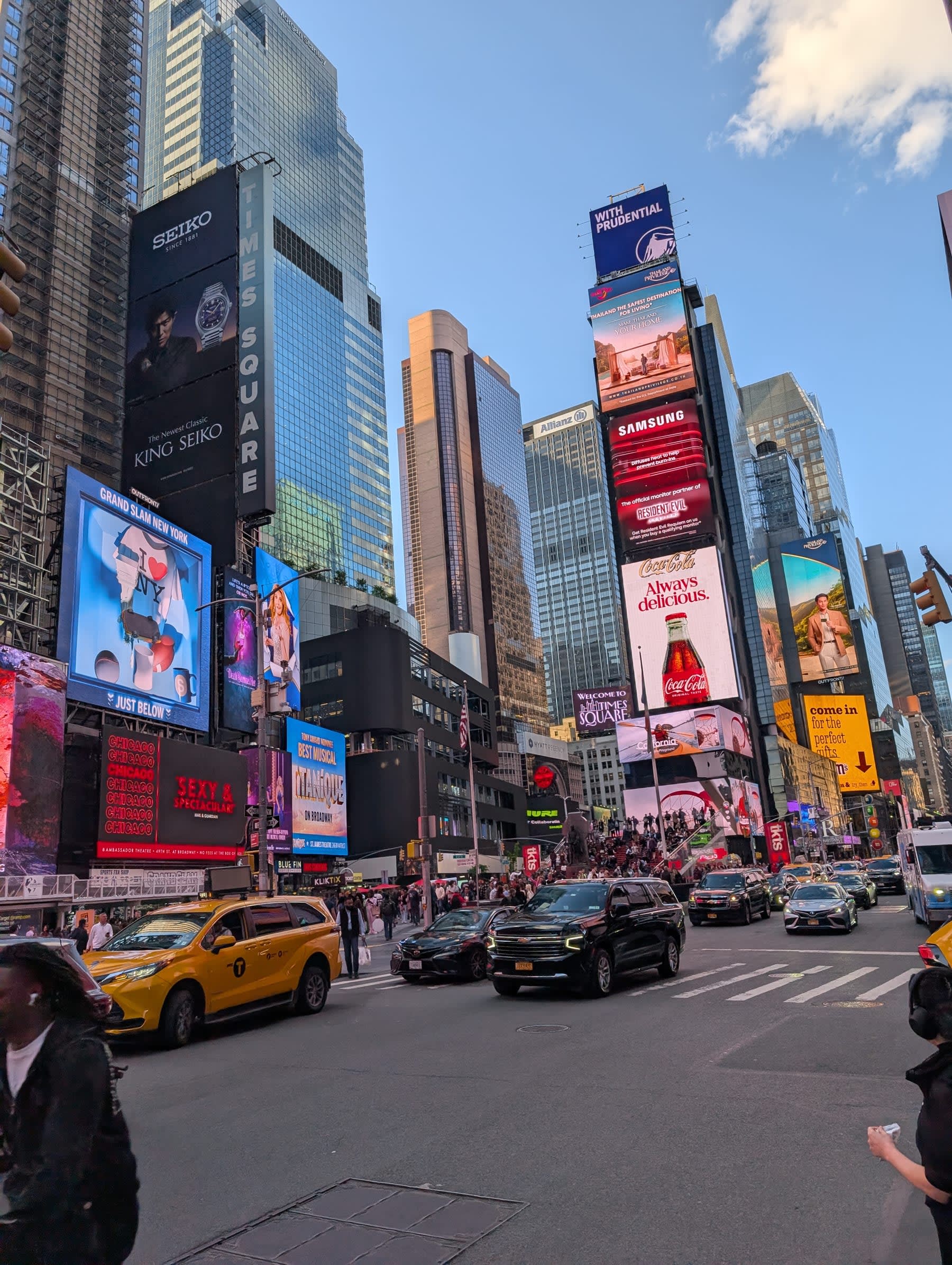 Times Square at dusk — the Seiko, Samsung, and Coca-Cola billboards catching the last sky light, yellow cabs in the street below, the screen towers blazing against a blue early-evening sky