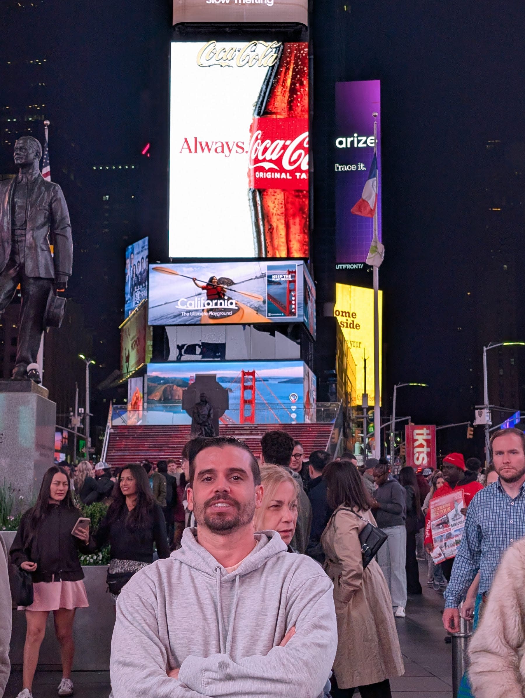 Pierre standing in Times Square at 2am — arms folded, the Coca-Cola "Always" billboard blazing directly behind him, a California tourism billboard visible, the square crowded with people who have decided that the evening is not finished yet