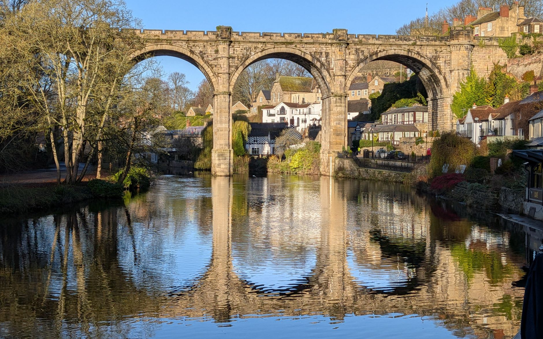 A wooden rowing boat on the calm green River Nidd beneath the tall arches of the railway viaduct