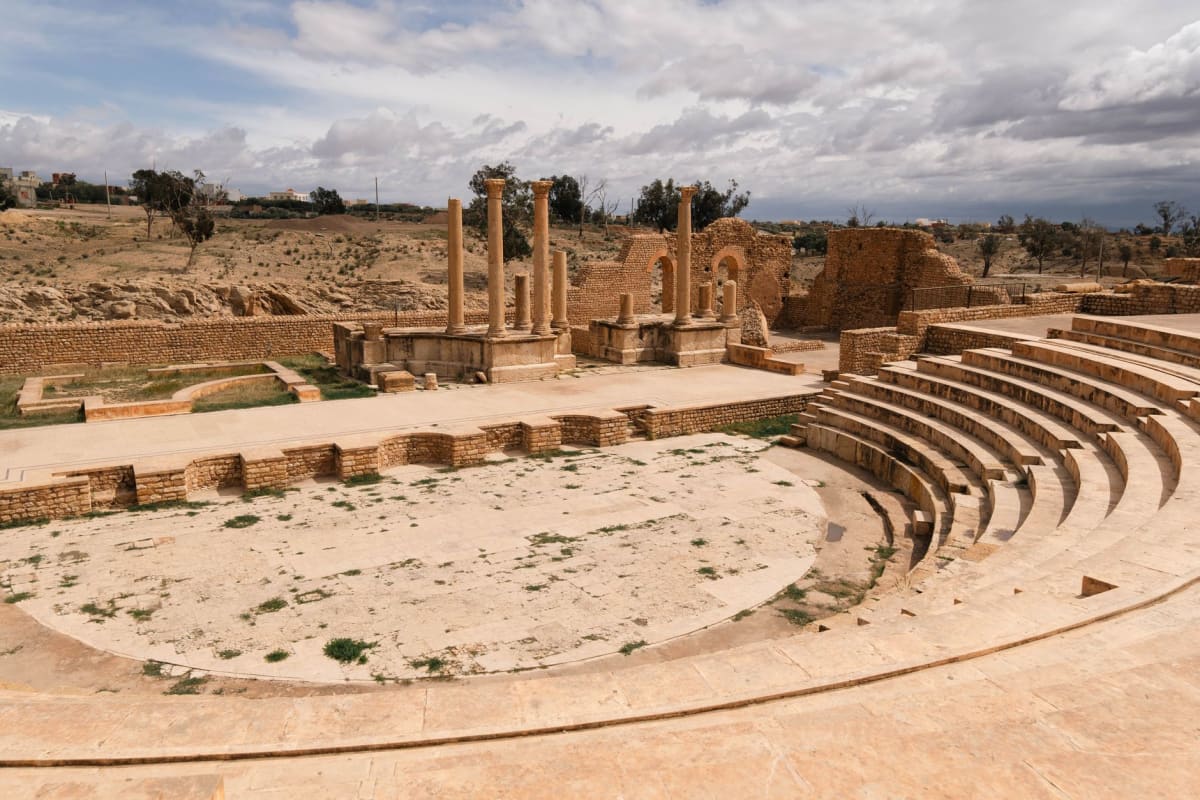 The Roman theater at Timgad seen from the upper seating tiers, the stage and backdrop visible against the mountain plateau