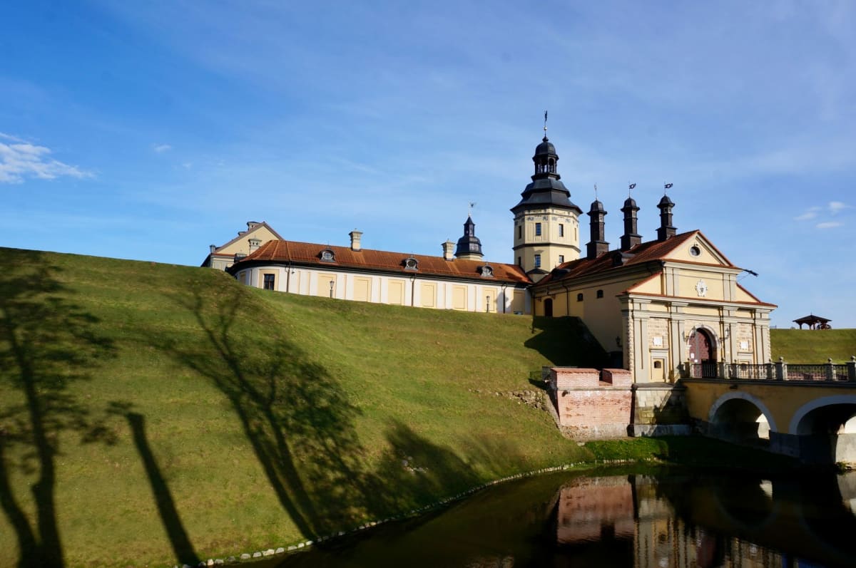 The outer walls and moat of Mir Castle seen from the road approach, the five towers rising against a pale Belarusian sky at dusk