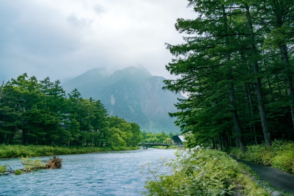 Le pont suspendu en bois Kappa-bashi enjambant la limpide rivière Azusa à Kamikōchi, des marcheurs le traversant avec les sommets d'Hotaka striés de neige derrière
