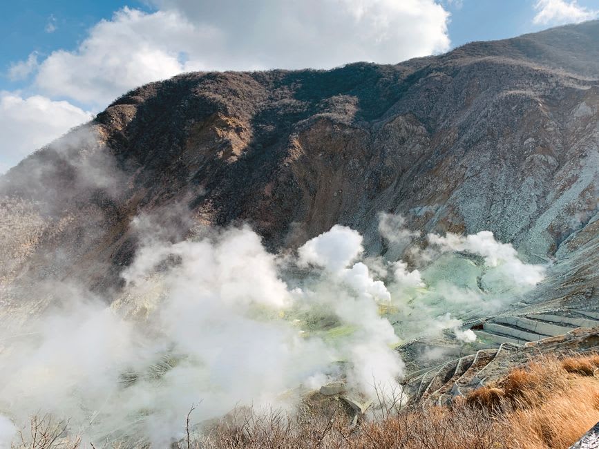 De la vapeur s'élevant d'un bain thermal extérieur à Noboribetsu au crépuscule, eau minérale laiteuse cernée de roche sombre tandis que la neige commence à tomber