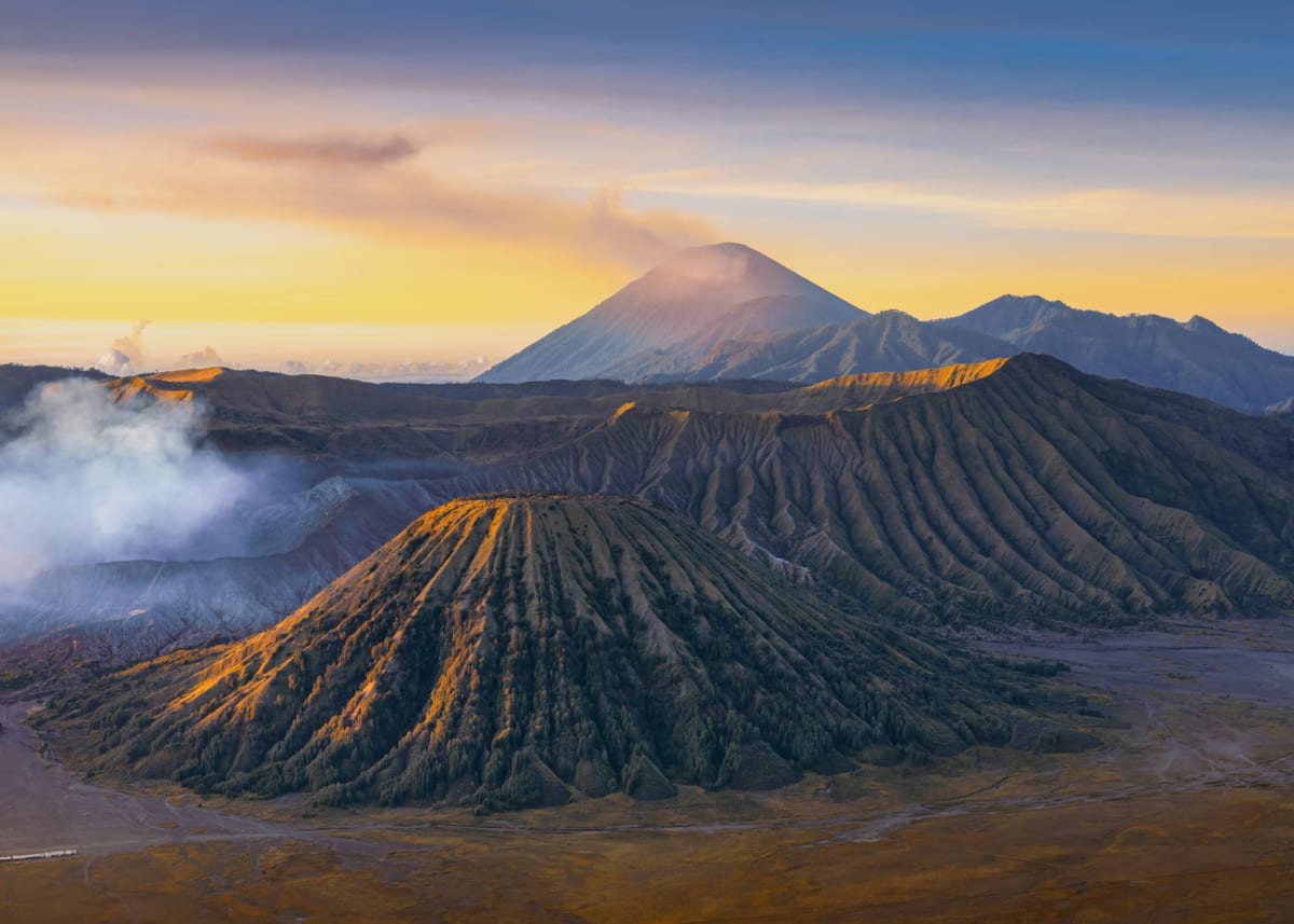 A Tenggerese pony handler and his horse silhouetted against Mount Bromo's smoking crater in the early morning light, the black sand stretching behind them