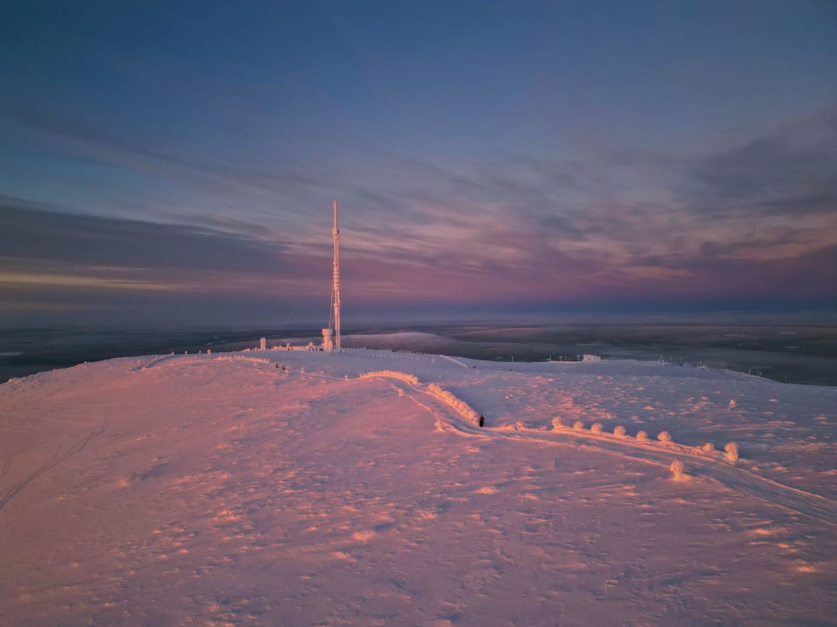 Una cabaña de sauna de leña en Luosto, pinos nevados rodeándola, una débil aurora sobre la línea de los árboles en el cielo nocturno
