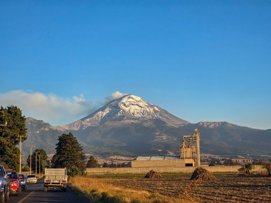 Terres agricoles plates et fertiles entourant Acatzingo à l'heure dorée, des rangées de maïs et des champs de légumes s'étendant vers la silhouette lointaine et floue des volcans, une route de terre rectiligne les traversant
