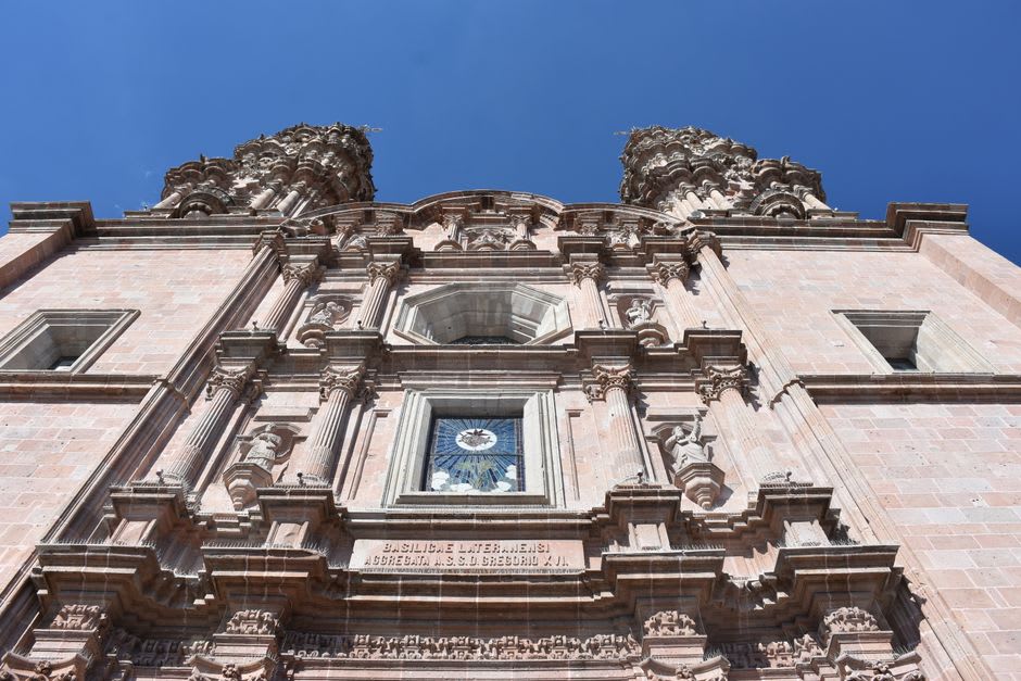 La Parroquia de Nuestra Señora de la Asunción al mediodía, su fachada barroca de dos torres proyectando largas sombras sobre el atrio de piedra vacío.