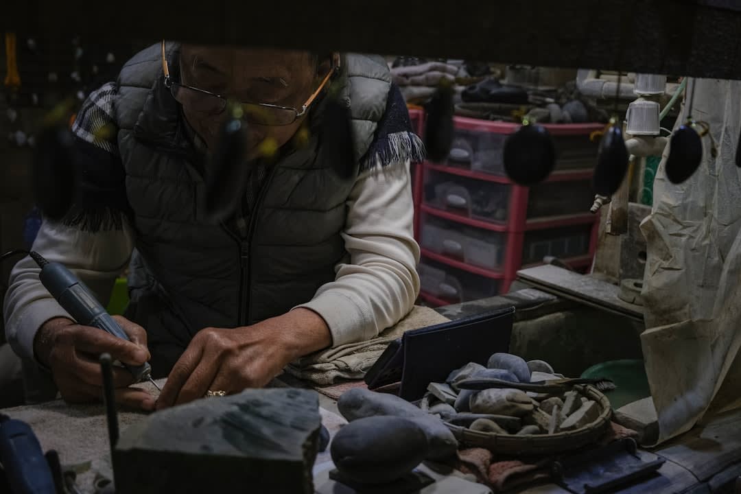 Un artisan de Cofradía de Suchitlán sculptant un masque de jaguar, sa gouge et son maillet visibles, des copeaux de bois frais sur le sol de l'atelier et des masques peints terminés suspendus au mur derrière lui