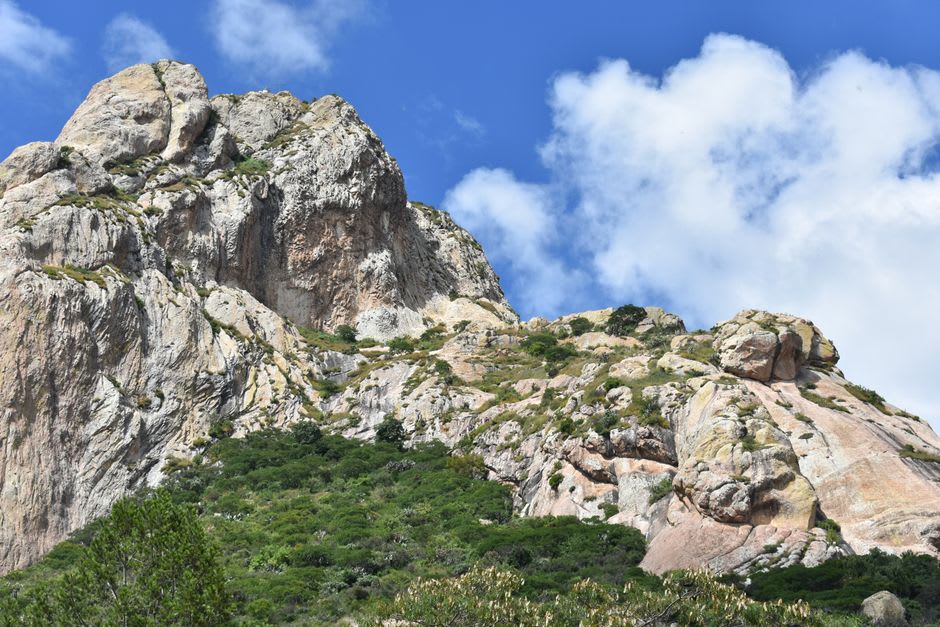 Randonneurs gravissant la section inférieure de la Peña de Bernal, la paroi de granit au-dessus d'eux révélant les stries géologiques d'une intrusion granitique vieille de 130 millions d'années, la vallée et le village visibles loin en contrebas