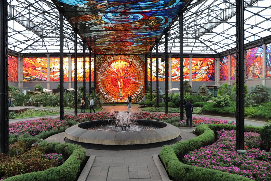 The interior of the Cosmovitral in Toluca, colored light from massive stained-glass panels falling across tropical plants and visitors below