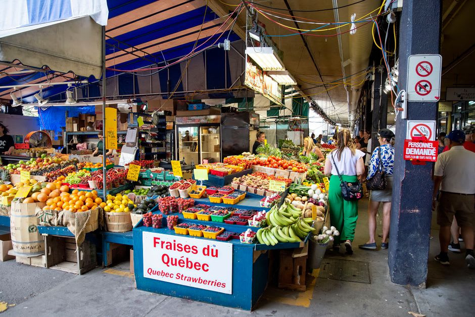 Un vendedor acomodando manojos de hierbas frescas y flores en un puesto del mercado Jean-Talon, multitud matutina pasando