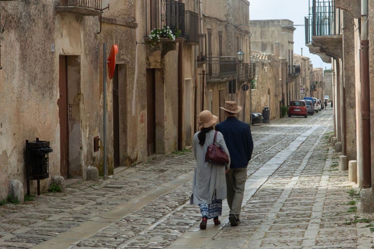 Empty grey cobbled lane in Erice winding between medieval stone houses, soft mist drifting through and a glimpse of a courtyard full of geraniums
