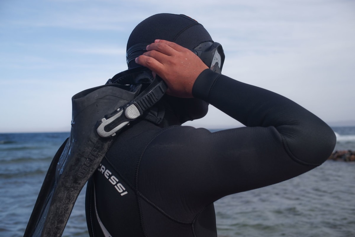 A haenyeo diver in a black wetsuit surfacing at the rocky Jeju coastline, floating on her back with diving fins visible, orange buoy nearby