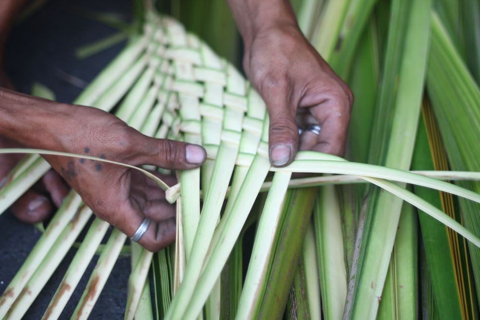Woman sitting outdoors weaving a patterned basket from pale palm fronds