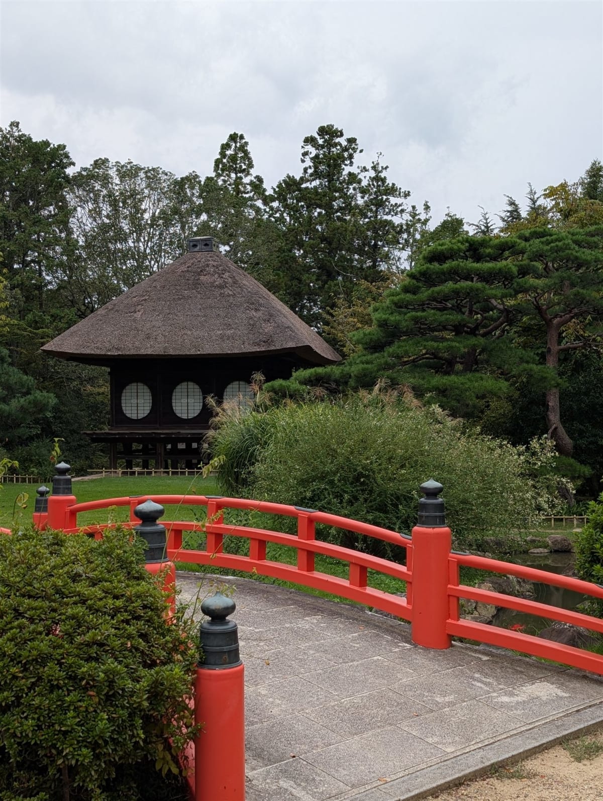 An arched vermilion bridge crossing a pond toward a thatched-roof pavilion in the Manyo Botanical Garden behind Kasuga Taisha