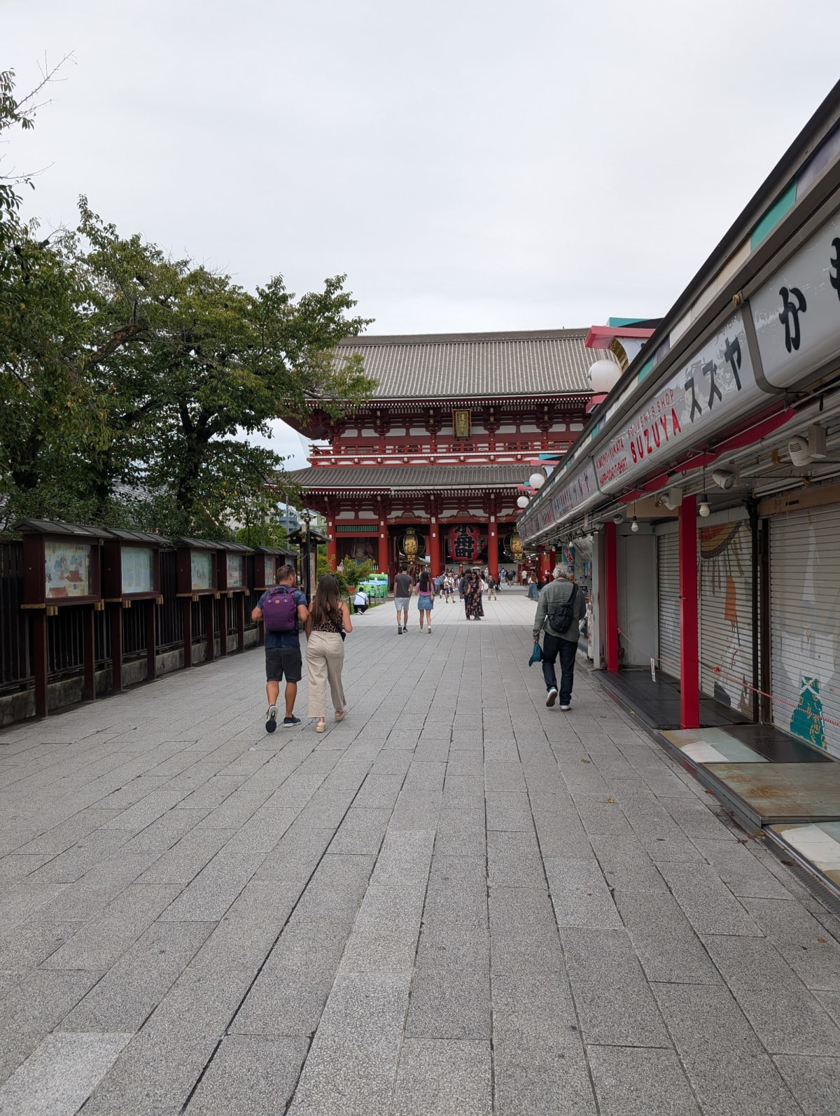 Lia and Pierre walking toward Senso-ji's inner gate at dusk, shuttered souvenir stalls lining the approach on either side