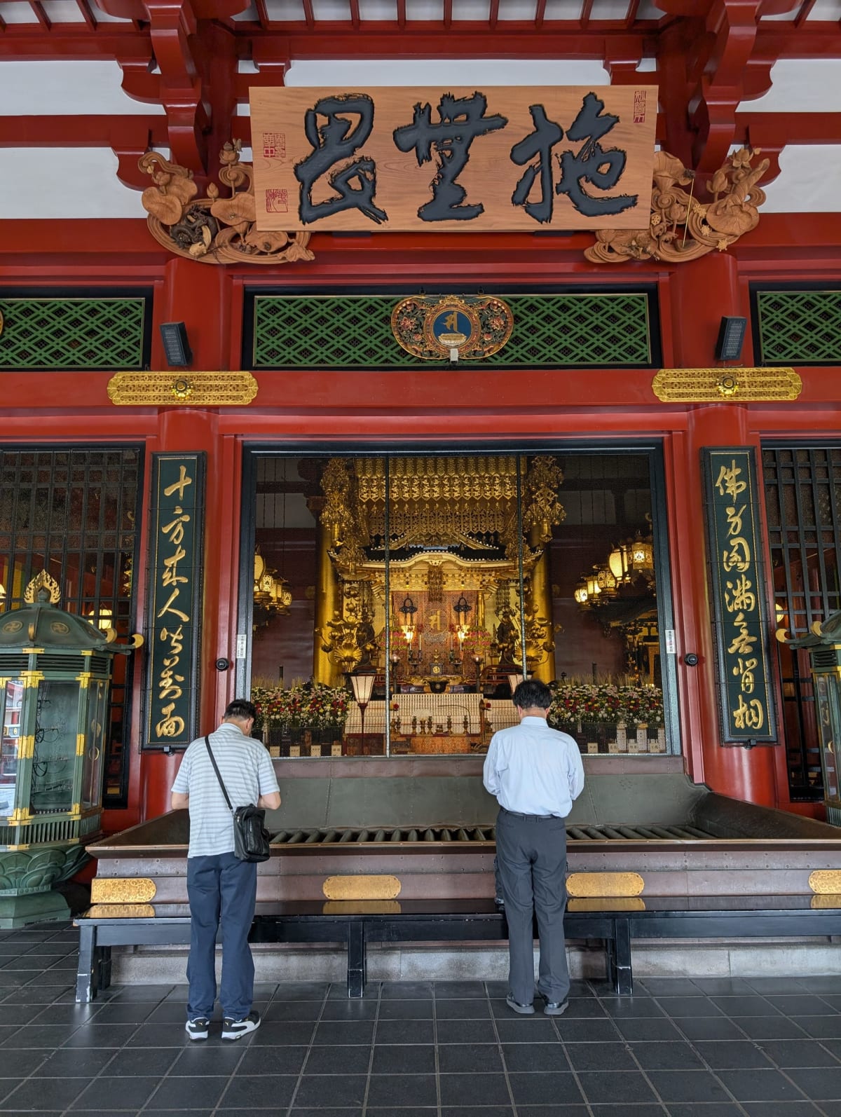 Two men standing at the altar rail inside Senso-ji's main hall, bowing in front of the illuminated golden inner shrine